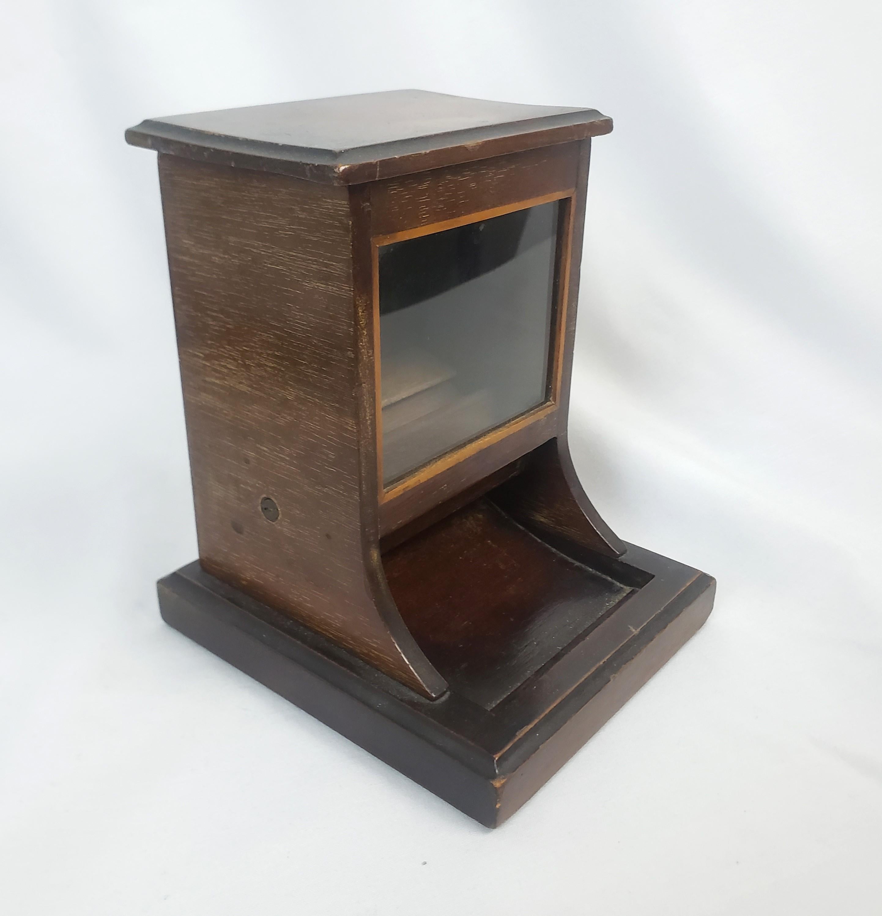 This antique table top cigarette dispenser was made by the renowned Dunhill firm of England and dates to approximately 1920 and done in the period Art Deco style. The dispenser is composed of wood with a glass front window and a slide closure top.