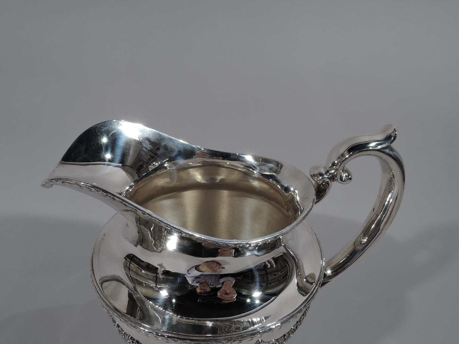 Regal sterling silver water pitcher in Empire pattern. Made by Durgin in Concord, New Hampshire. Ovoid with helmet mouth, leaf-capped scroll handle, and round foot. Applied ornament: Two oval ribbon-tied imbricated-leaf frames surmounted by crown,