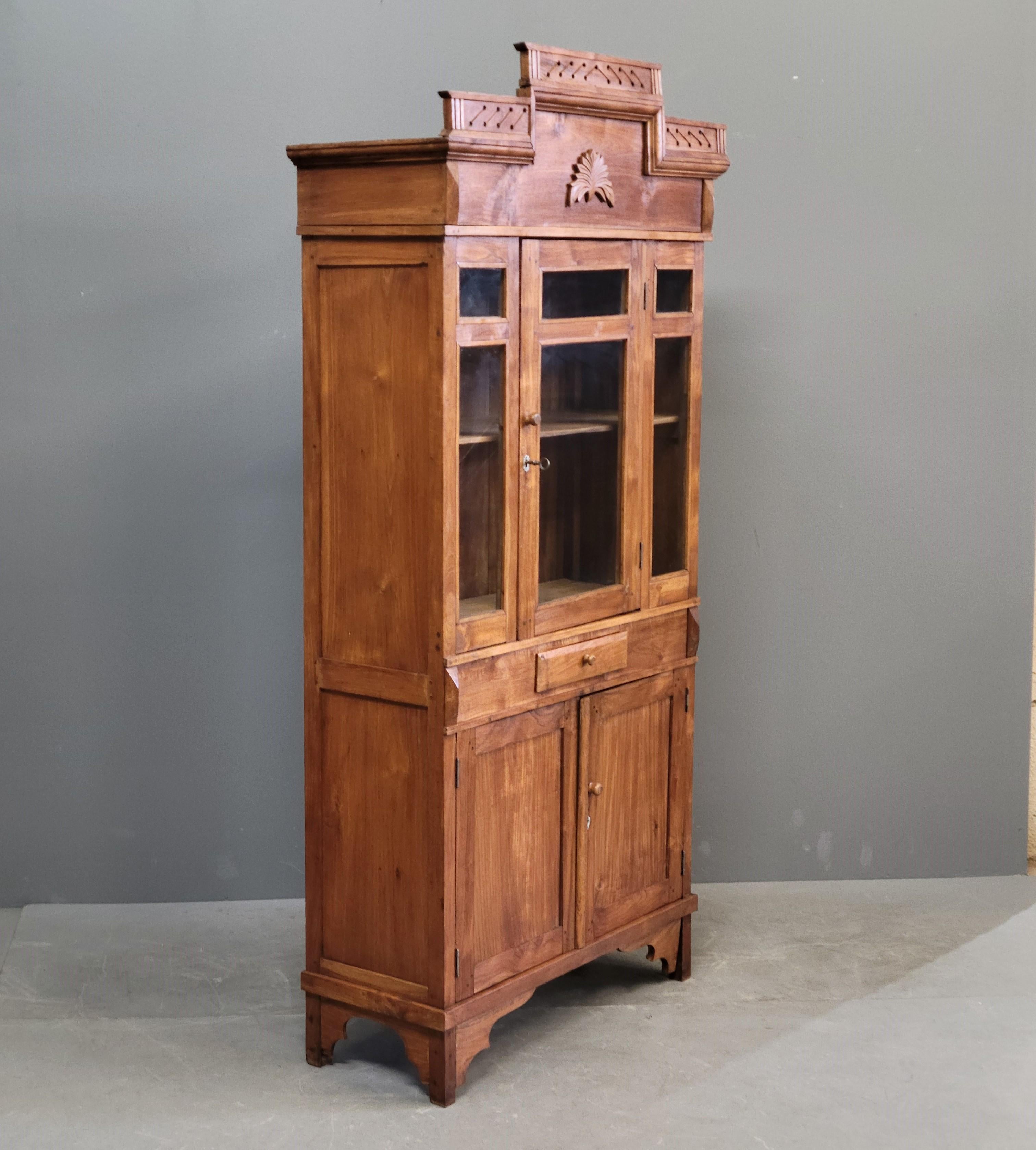 A charming teak Dutch Colonial storage cabinet with glass doors from Java, Indonesia. The stepped crown with applied carving is distinctly Dutch. Ample storage below, storage drawer, and glass door storage above for display space. Imperfections in