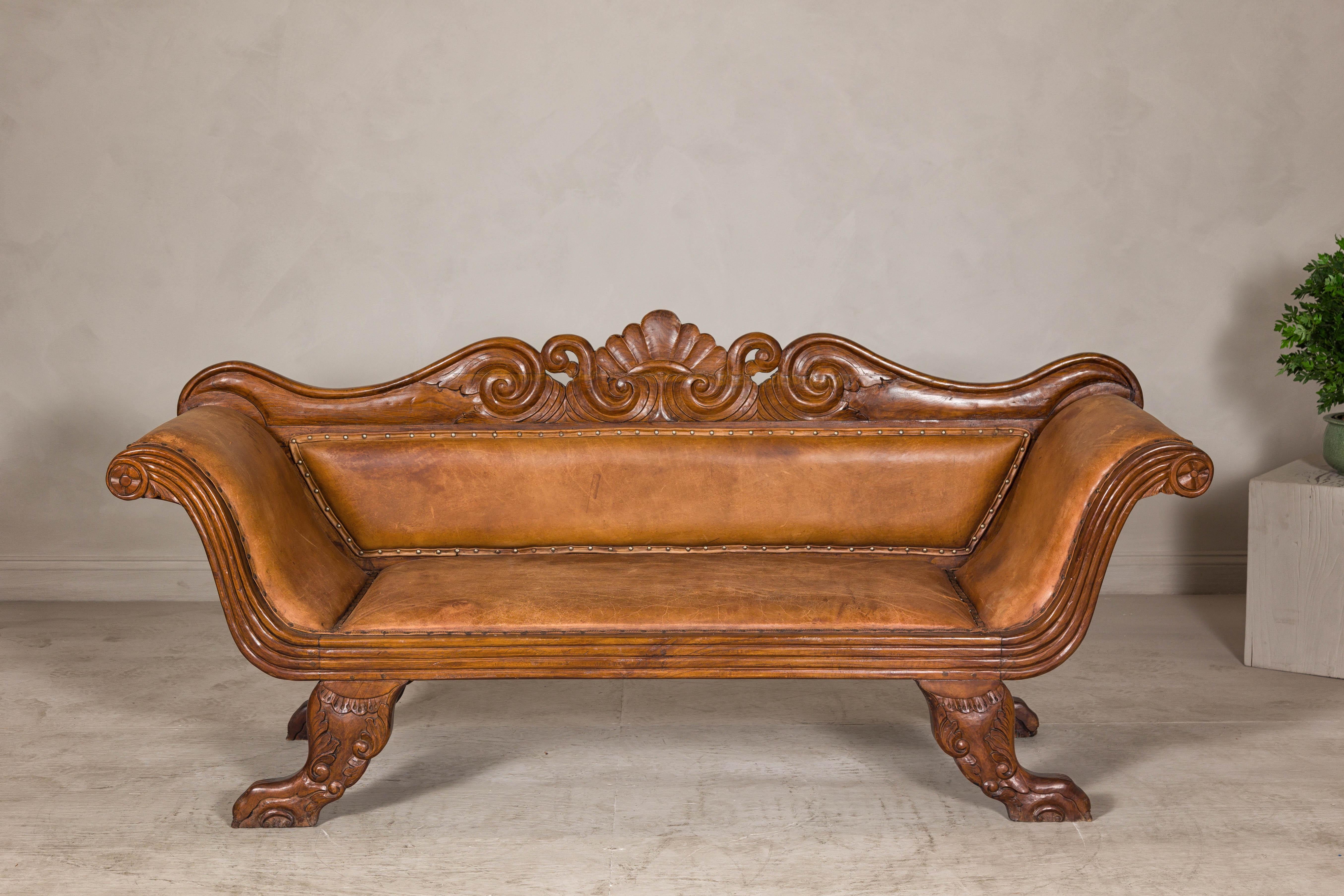 A Dutch Colonial teak wood settee from circa 1880-1920 with original brown leather and carved motifs. This exquisite Dutch Colonial settee showcases the timeless elegance of antique craftsmanship with its carved wood frame and original leather