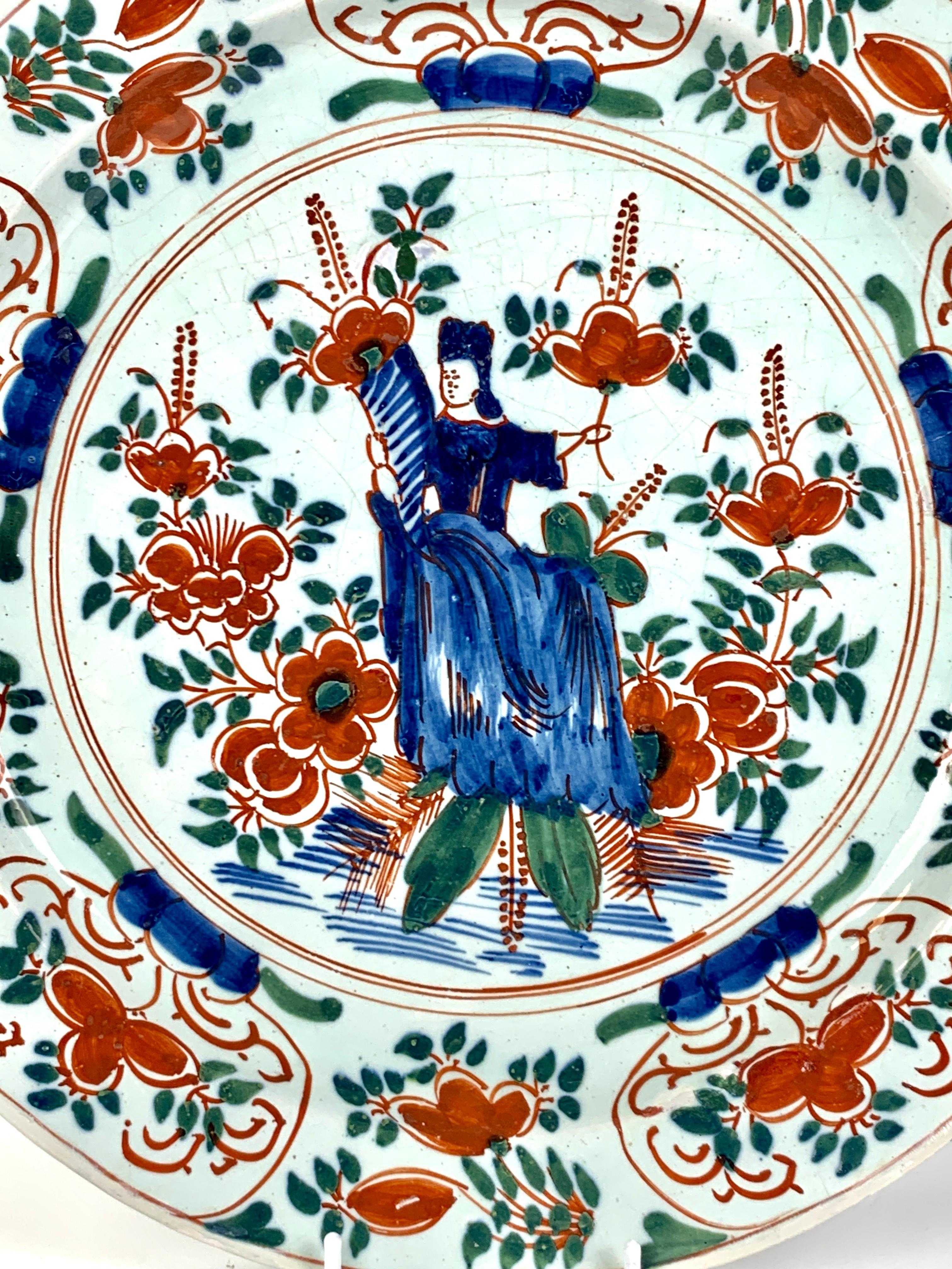 This lovely Delft charger shows a hand-painted scene with a seated young lady holding a horn of plenty. 
Dressed in cobalt blue, she is surrounded by iron red flowers with forest green leaves.
The image is a traditional joyous symbol of harvest and