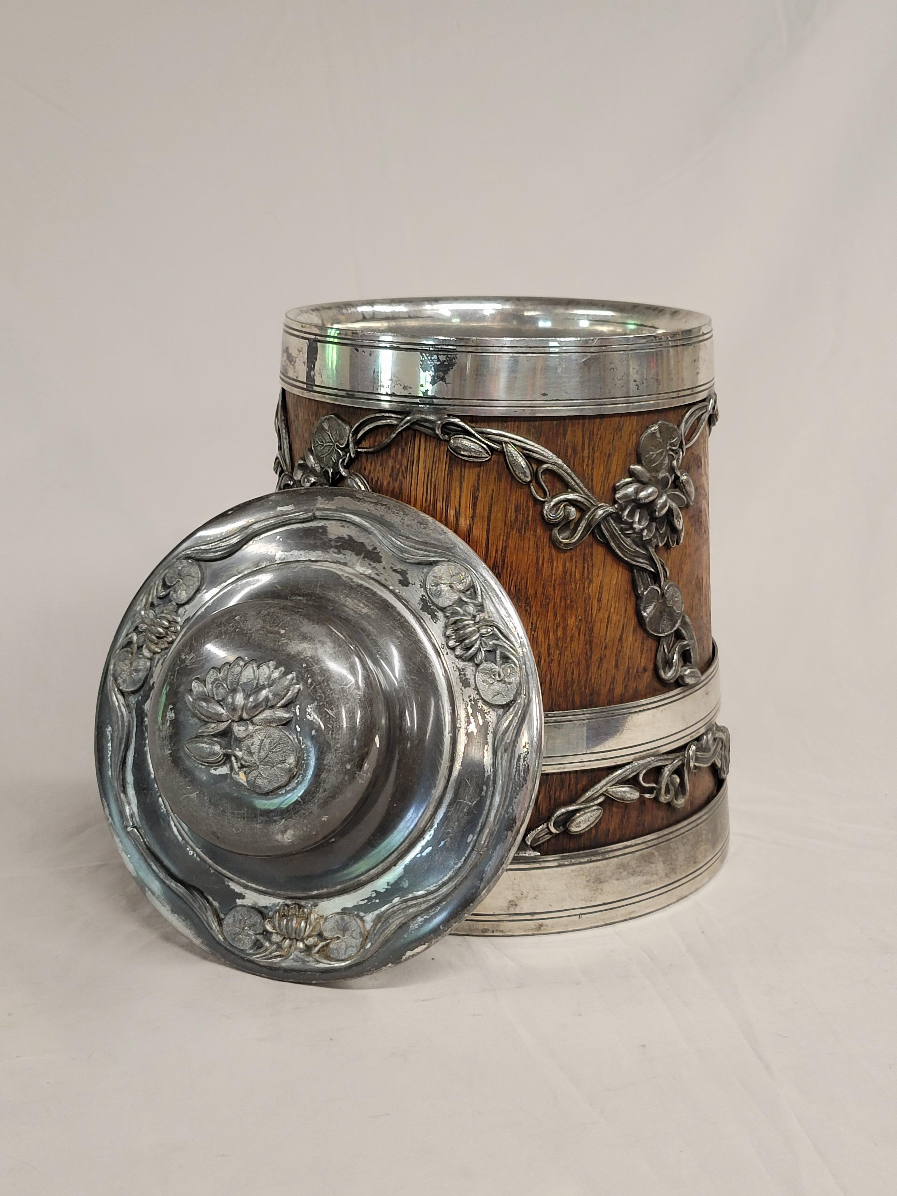 This elegant antique ice bucket was crafted by the St. Louis Silver Company, which was active from 1904-1912. The body was made of dense Flemish oak and ornamented with gorgeous art nouveau style water lilies in quadruple plate silver. St. Louis