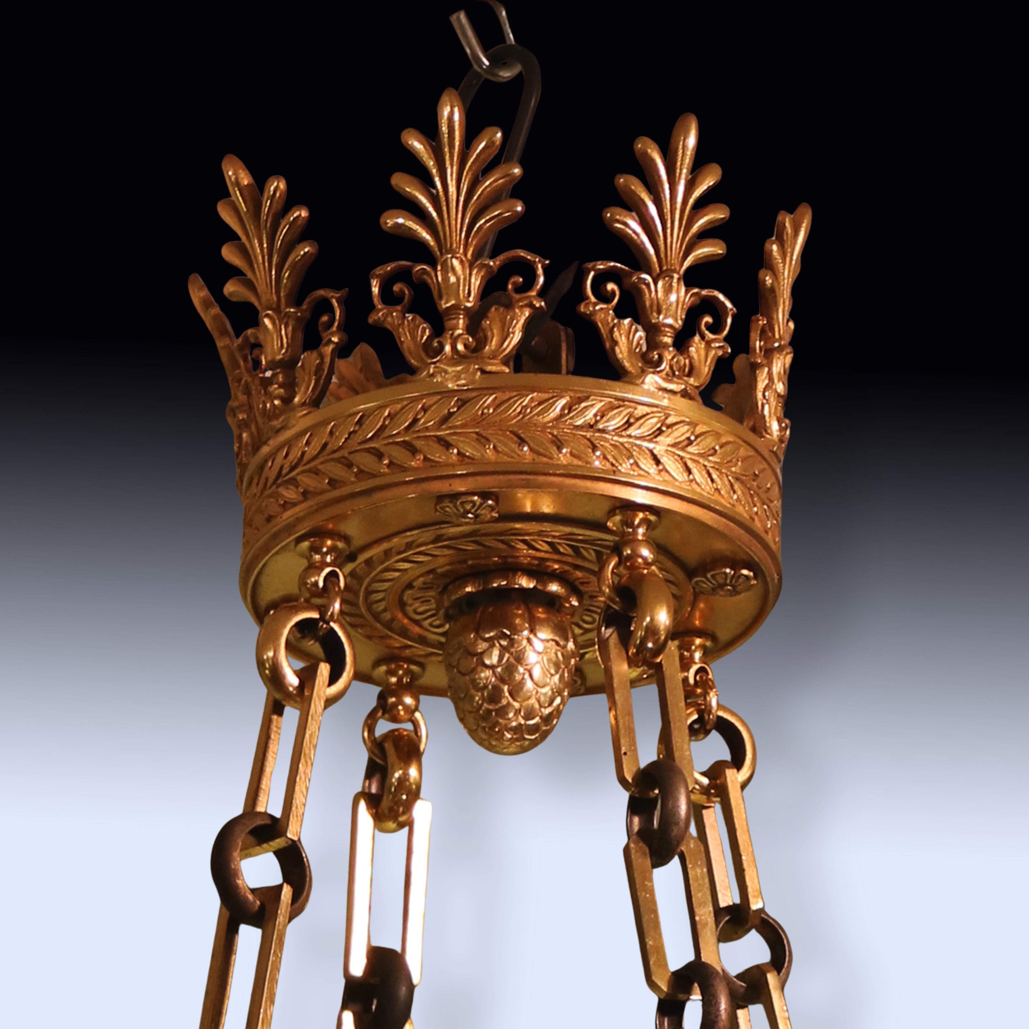 Lámpara de araña de 12 luces de estilo Imperio de principios del siglo XX, de bronce patinado verde y ormolina, con corona de antemuro y remate, que sostiene 4 cadenas decorativas.  La pieza tiene un flambeado central de cristal opaco y brazos de
