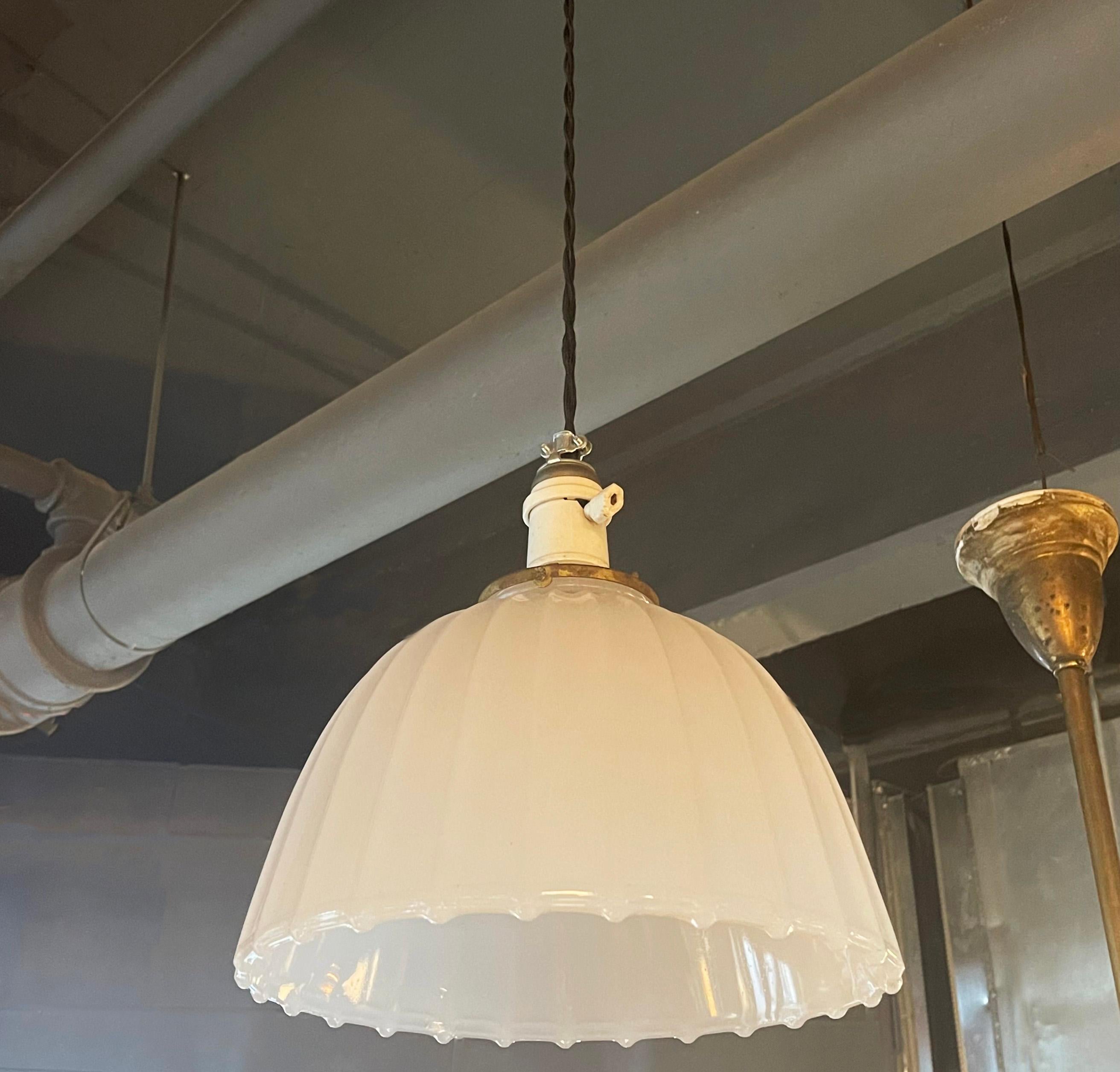 This elegant, early 20th century pendant light features a 10 inch height ribbed milk glass dome shade with scalloped edge paired with a white porcelain socket with paddle switch and brass fitter. The pendant is newly wired with 40 inches of black
