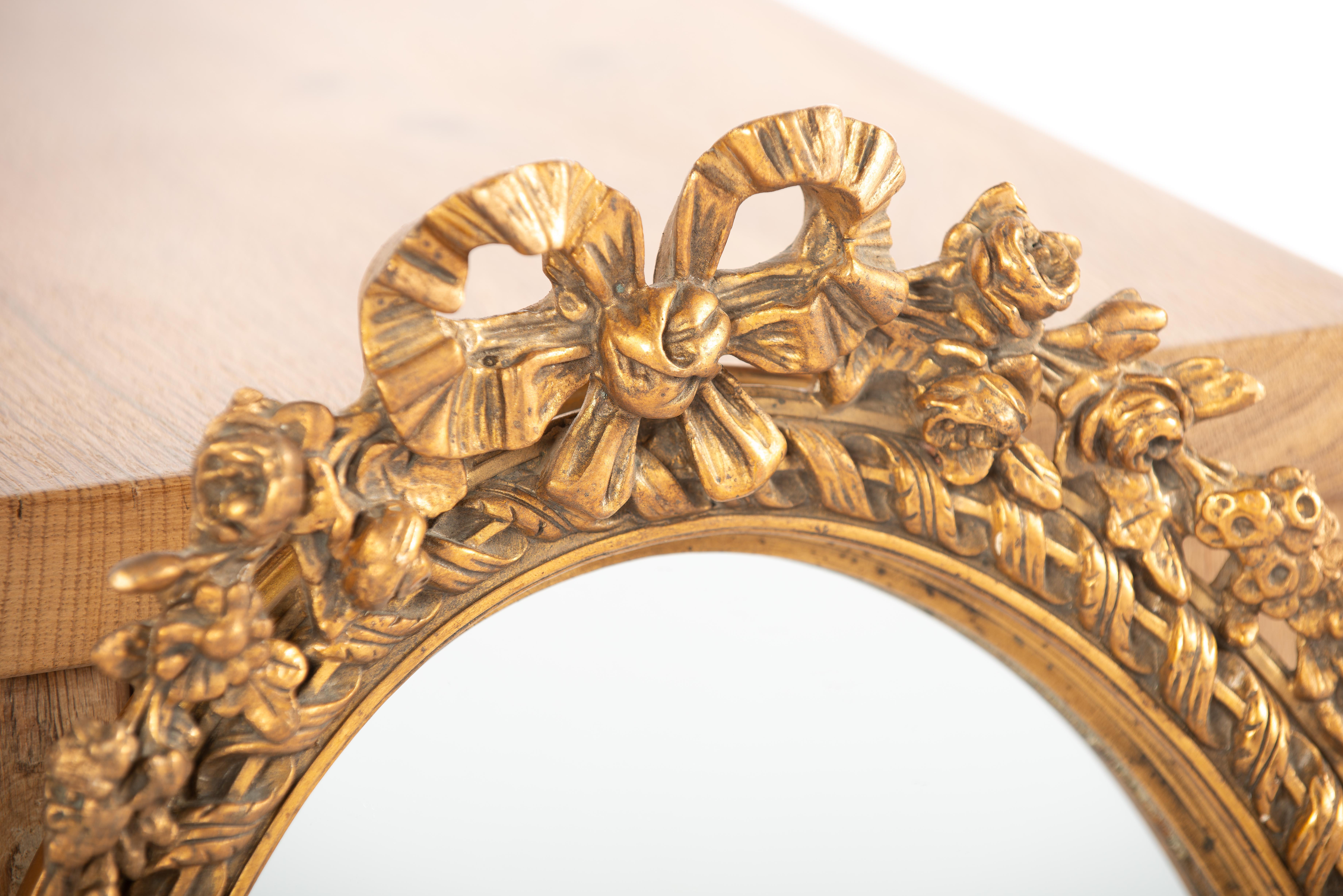 This is a beautiful small oval mirror that was crafted in France in the early 20th century, around 1910. The mirror features a frame made of lime wood and is adorned with plaster molding and ornaments. The top ornament consists of a tied ribbon