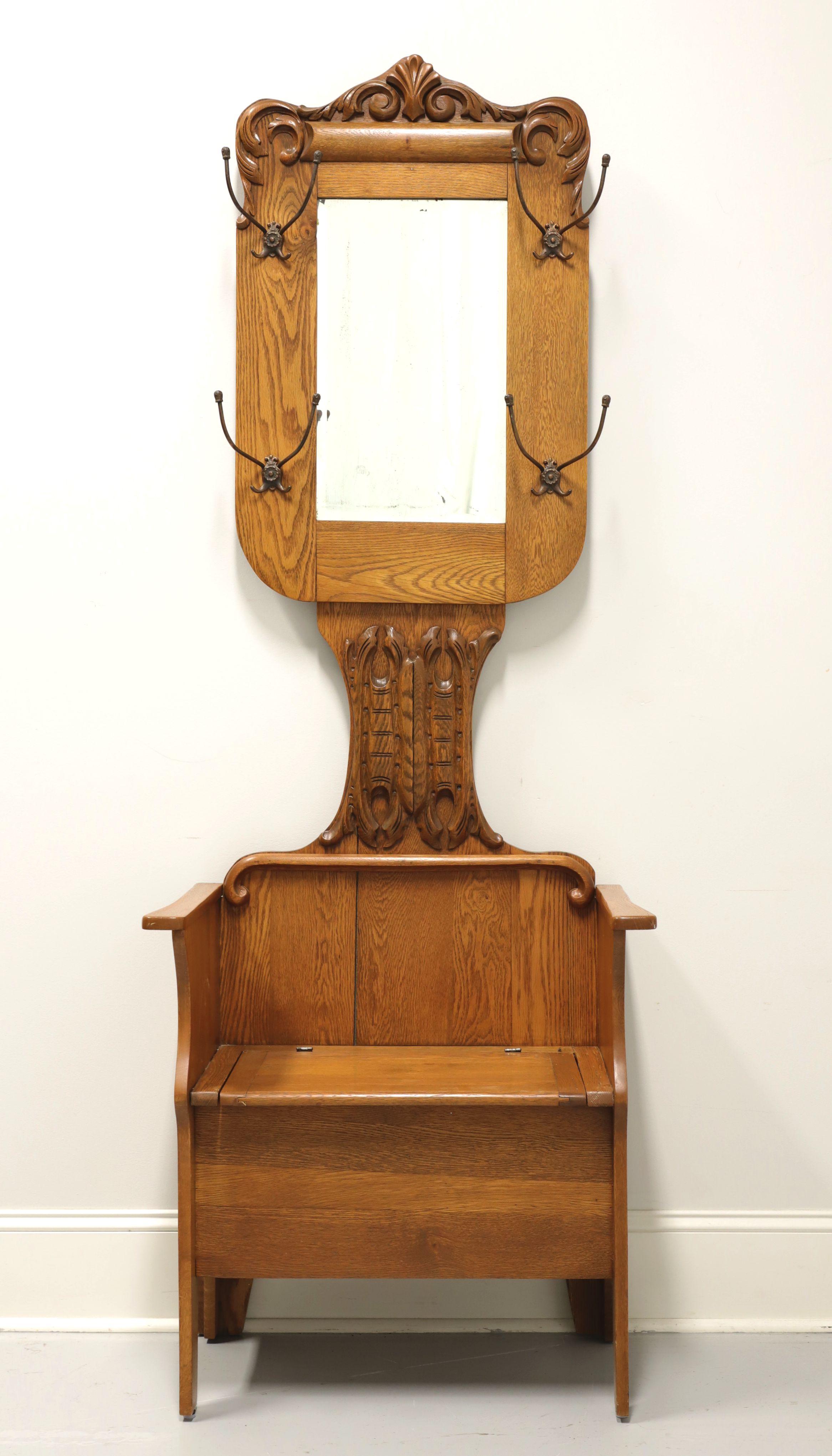 An antique Victorian style hall tree with mirror and bench, unbranded. Solid oak with brass hooks and mirror. Features decoratively carved framed mirror at top with garment hooks, decoratively carved center mirror support, lower bench with arms and
