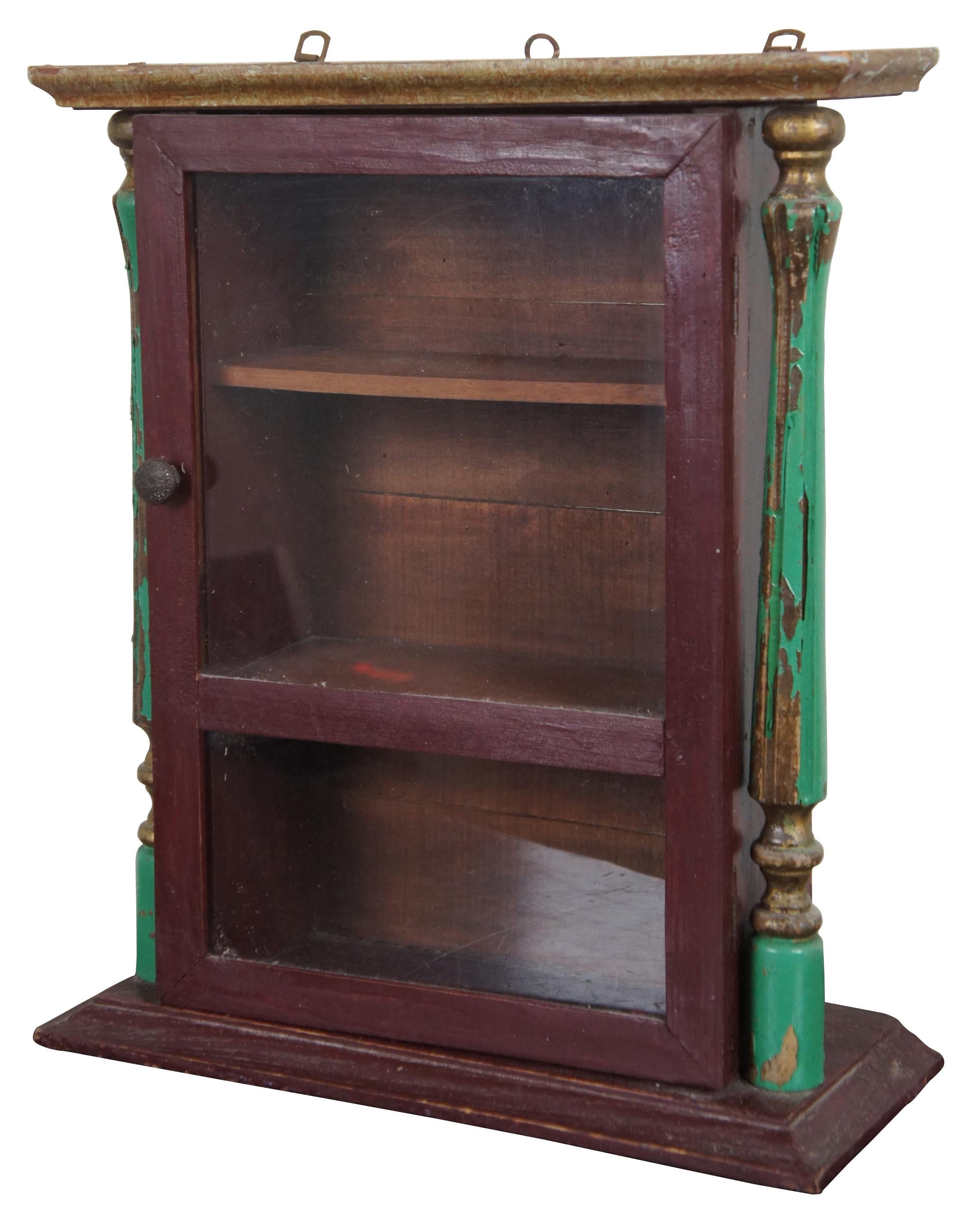 Primitive antique wooden wall cabinet painted dark red with a glass door and three shelves flanked by turned green painted columns. One side of the cabinet also sports an Art Deco floral motif. Measure: 18