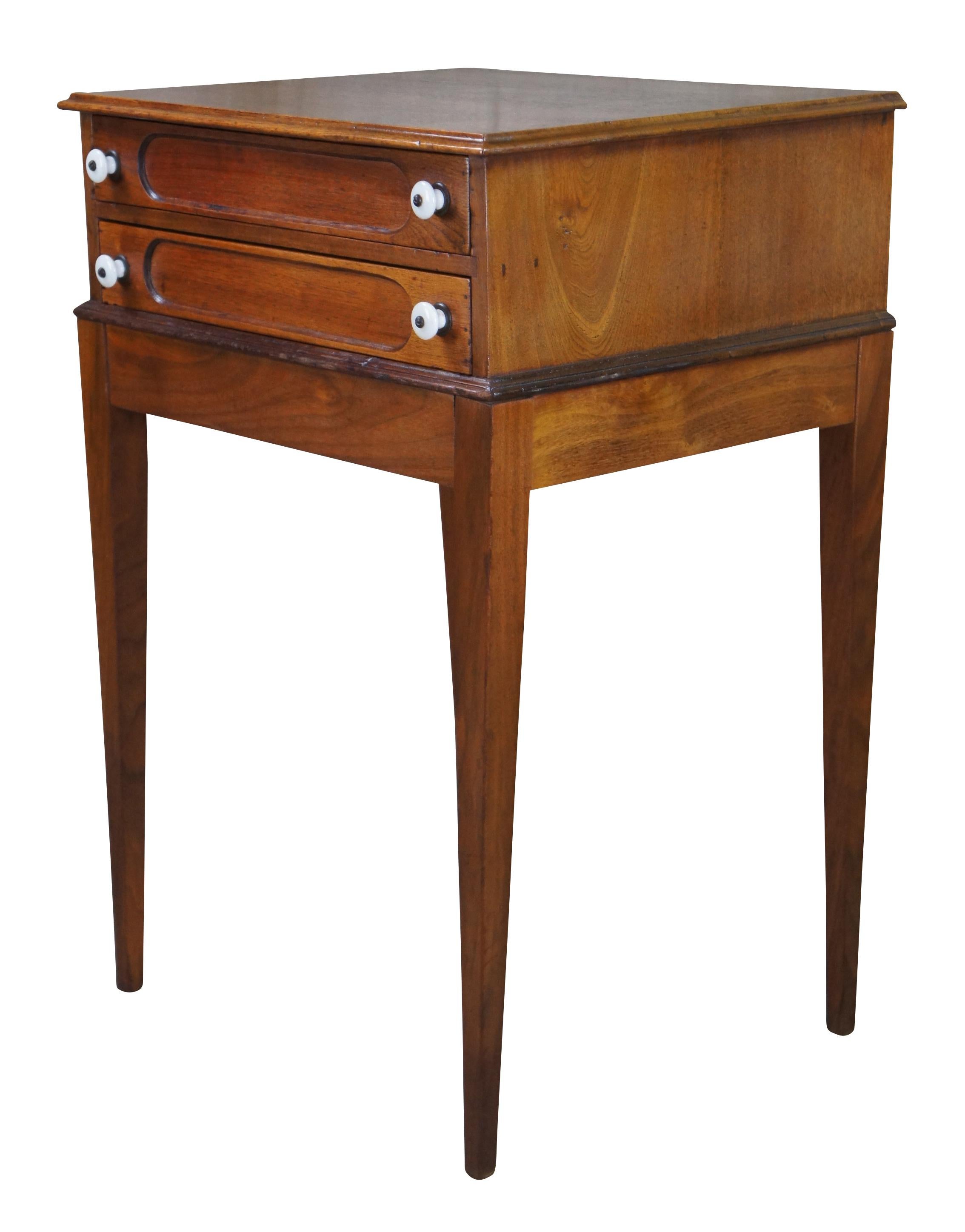 Antique early American spool cabinet side table. Made of walnut featuring two drawers with classic inset paneled fronts and porcelain knobs supported by square tapered legs. Measures: 28