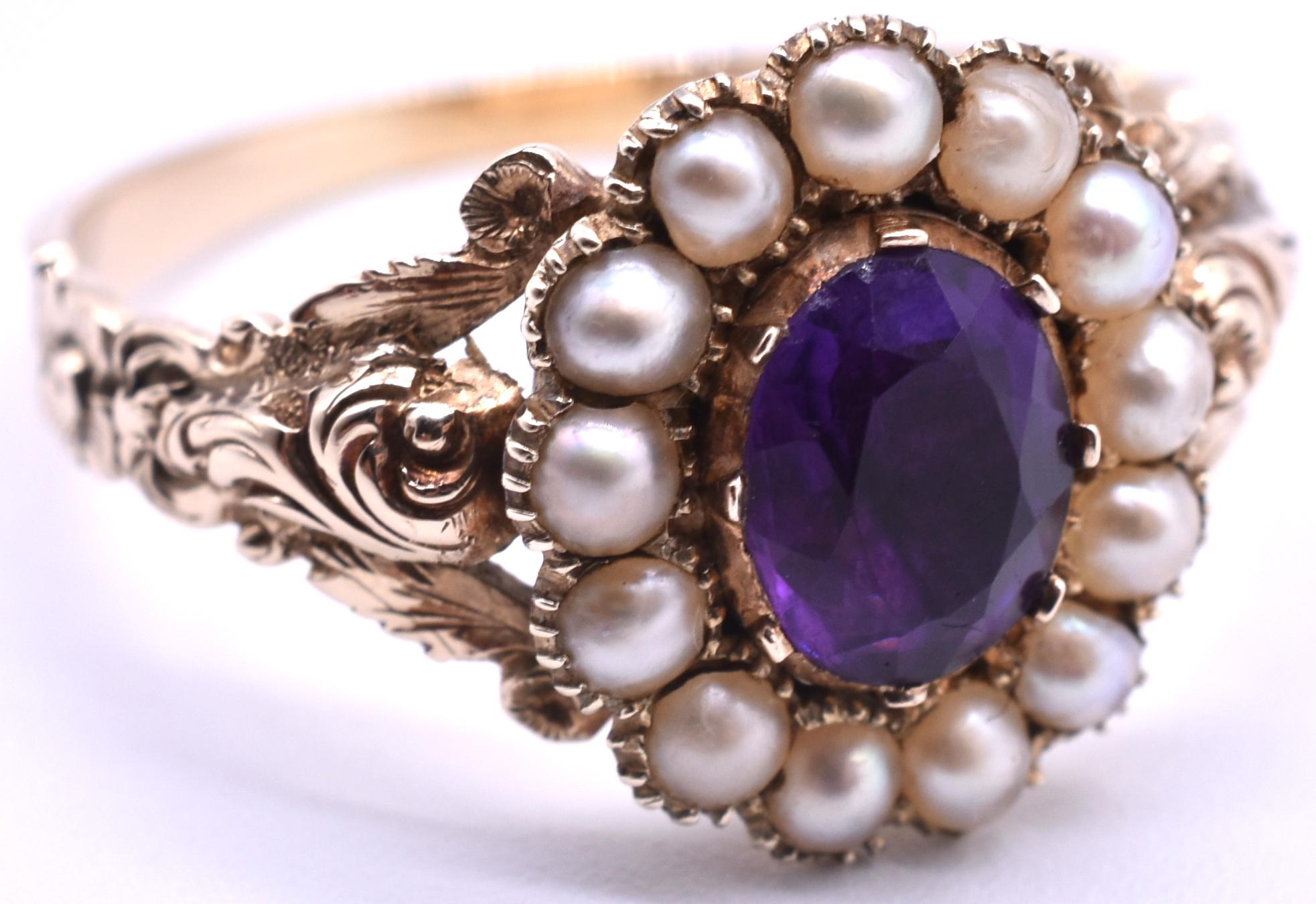 15k amethyst and seed pearl cluster ring with thirteen seed pearls bordering a beautiful amethyst oval stone. Repousse whorls adorn the shoulders and terminate in a small gold flower form. The setting is a foiled, closed back, 1850. The ring is US