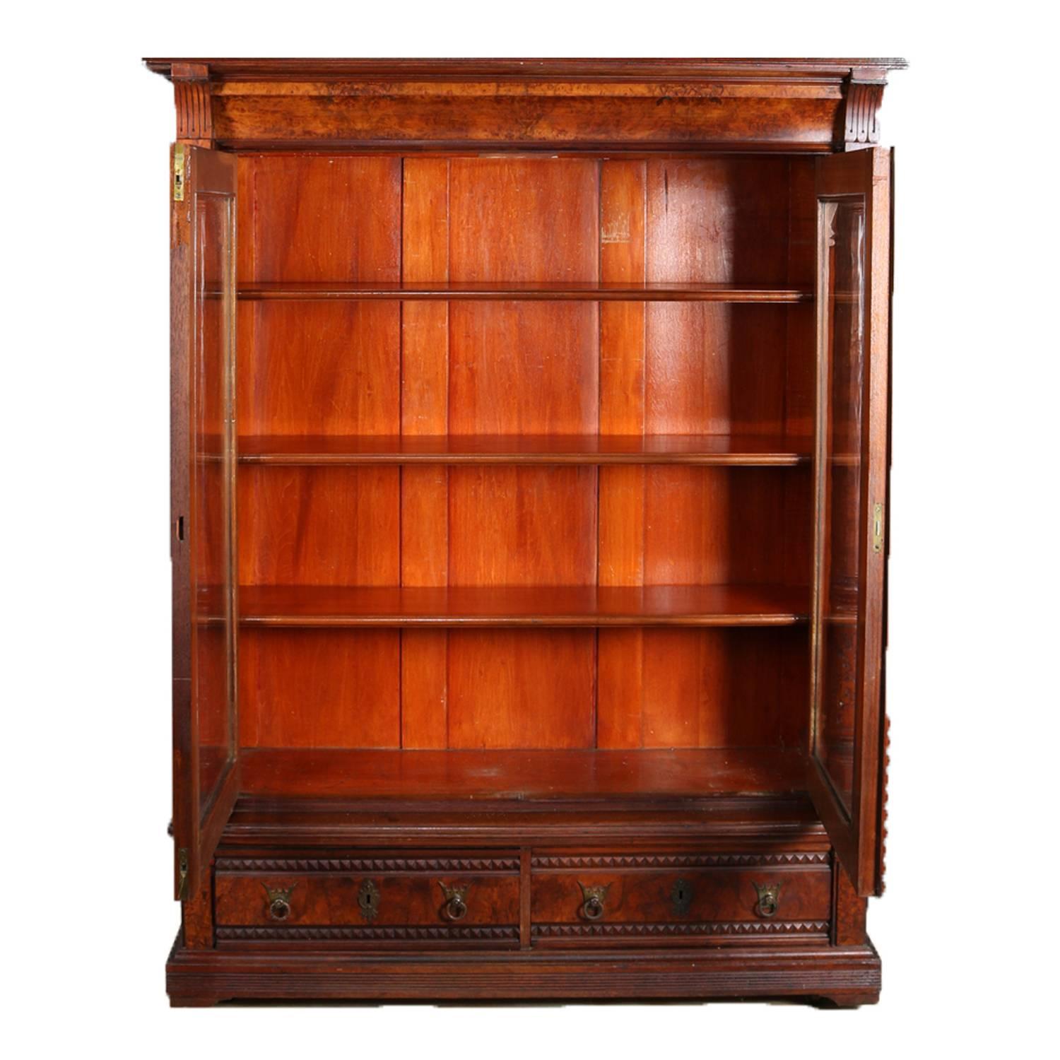 Antique Eastlake bookcase features burl walnut construction with brass gallery with cove molded crest above case with two glass doors opening to shelved interior flanked by floral and foliate incised columns and over two drawers with bronze crown