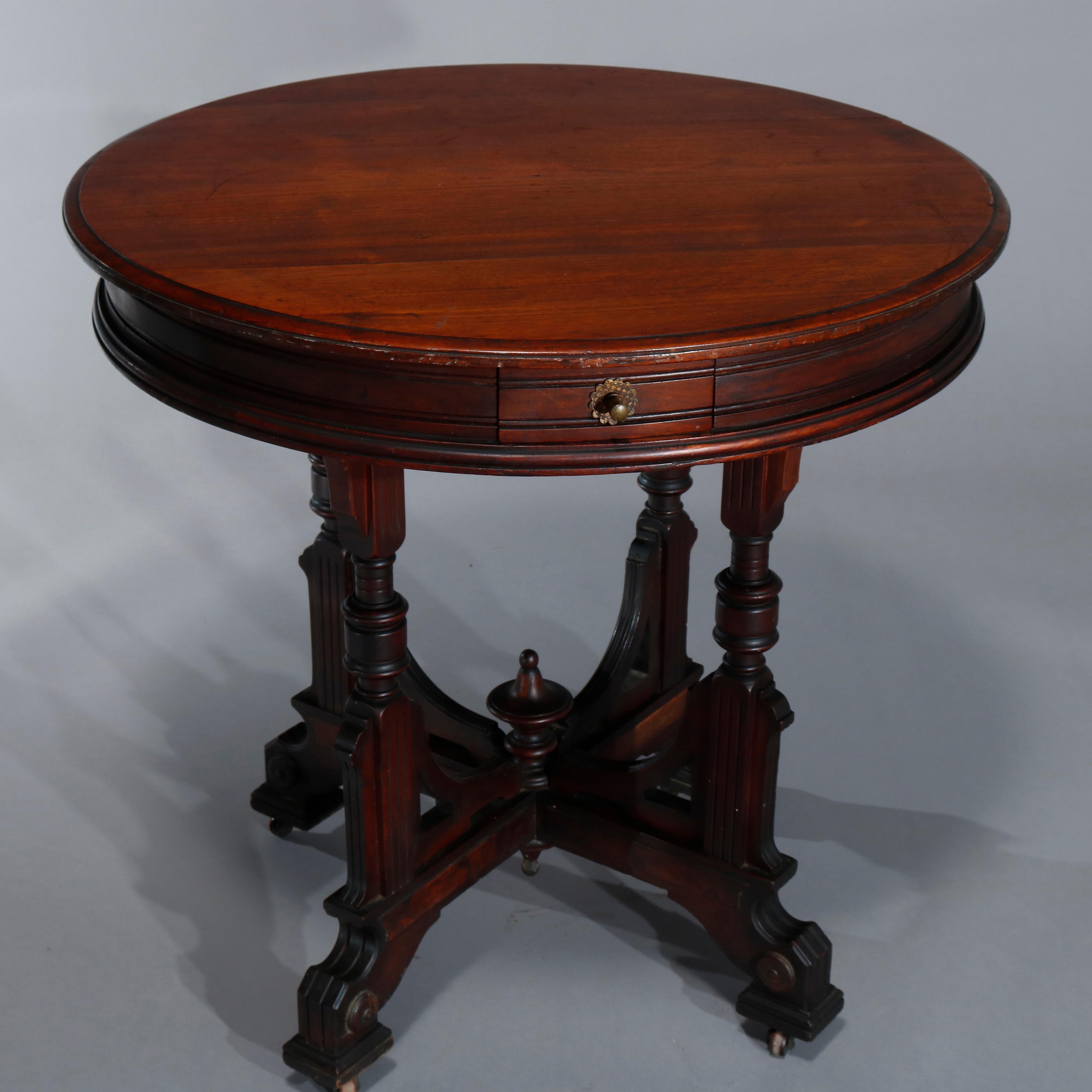An antique Eastlake parlor lamp table offers walnut construction with beveled top having deep skirt with single drawer raised on four carved legs having stretcher with corbels, central finials and terminating in stylized paw feet with casters, circa