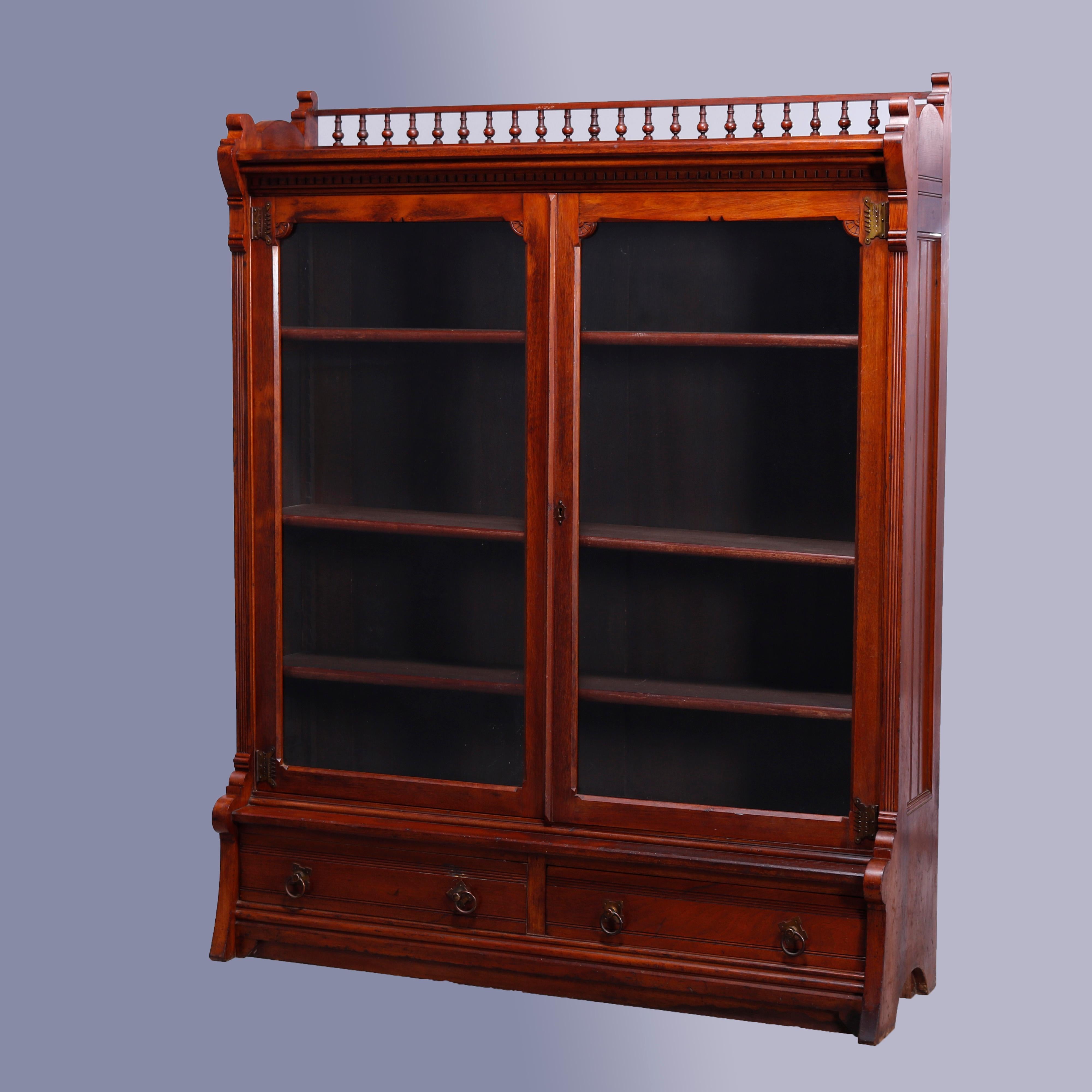 An antique Eastlake enclosed bookcase offers walnut construction with upper gallery having spindle rail and surmounting case with double glass doors opening to shelved interior and flanked by reeded column supports over double drawer base with