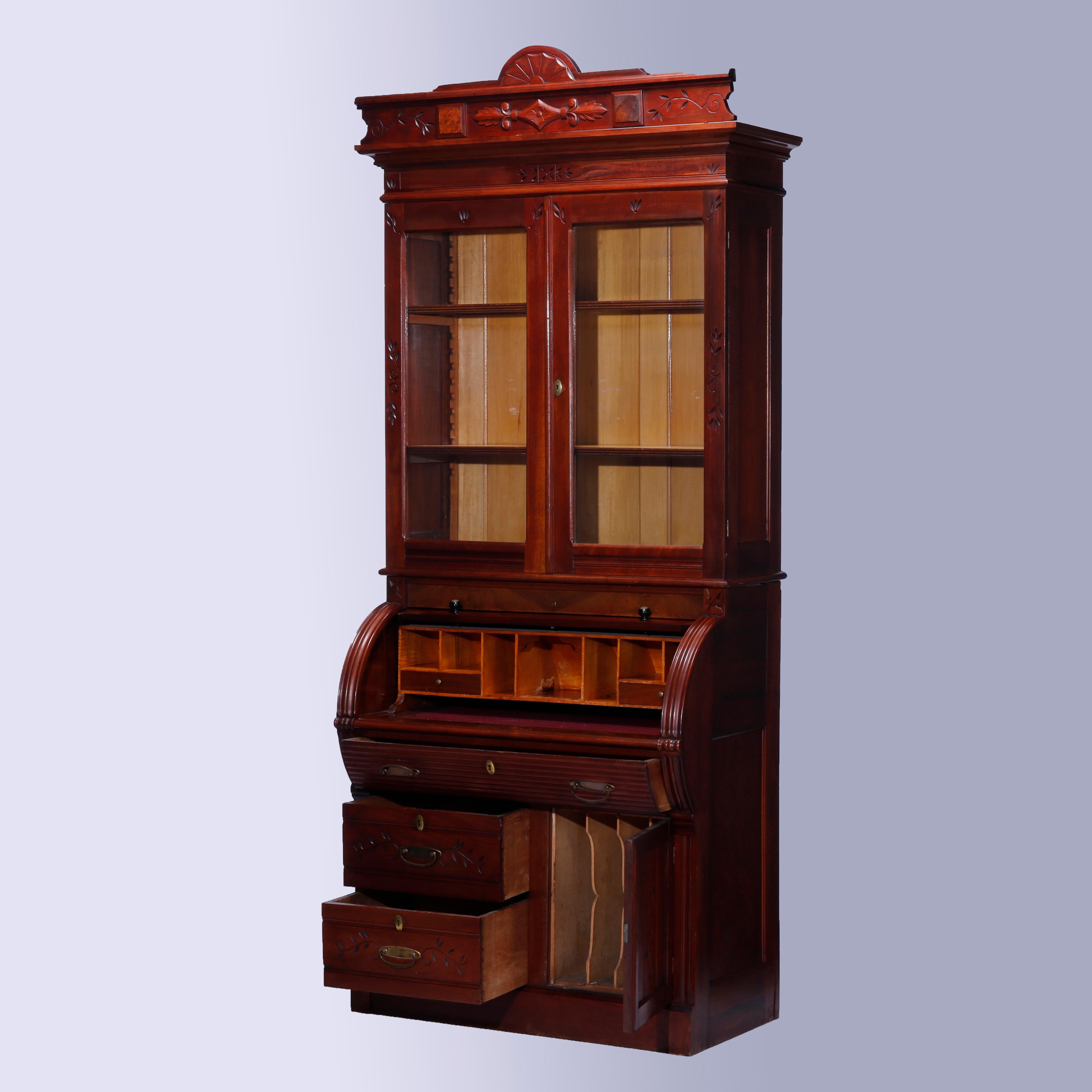 An antique Eastlake secretary offers walnut construction with carved sunburst crest over double glass door bookcase with adjustable shelves surmounting barrel roll desk opening to pull-out writing surface and pigeon holes storage compartments over