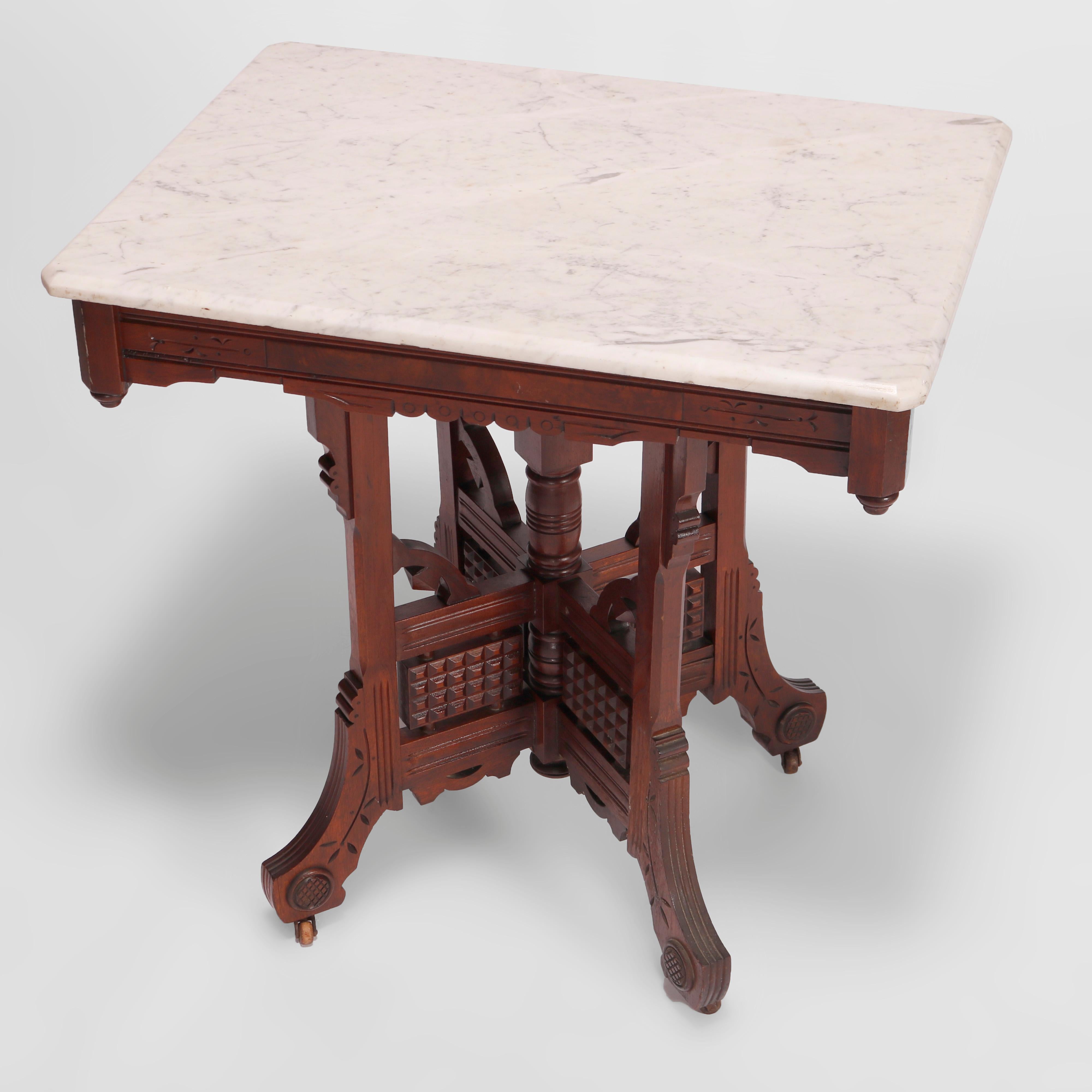 An antique Eastlake parlor table offers beveled clip-corner marble top over walnut base having burl insets, shaped skirt with drop finials, four supports with central column, raised on legs having incised foliate decoration and stylized scroll form