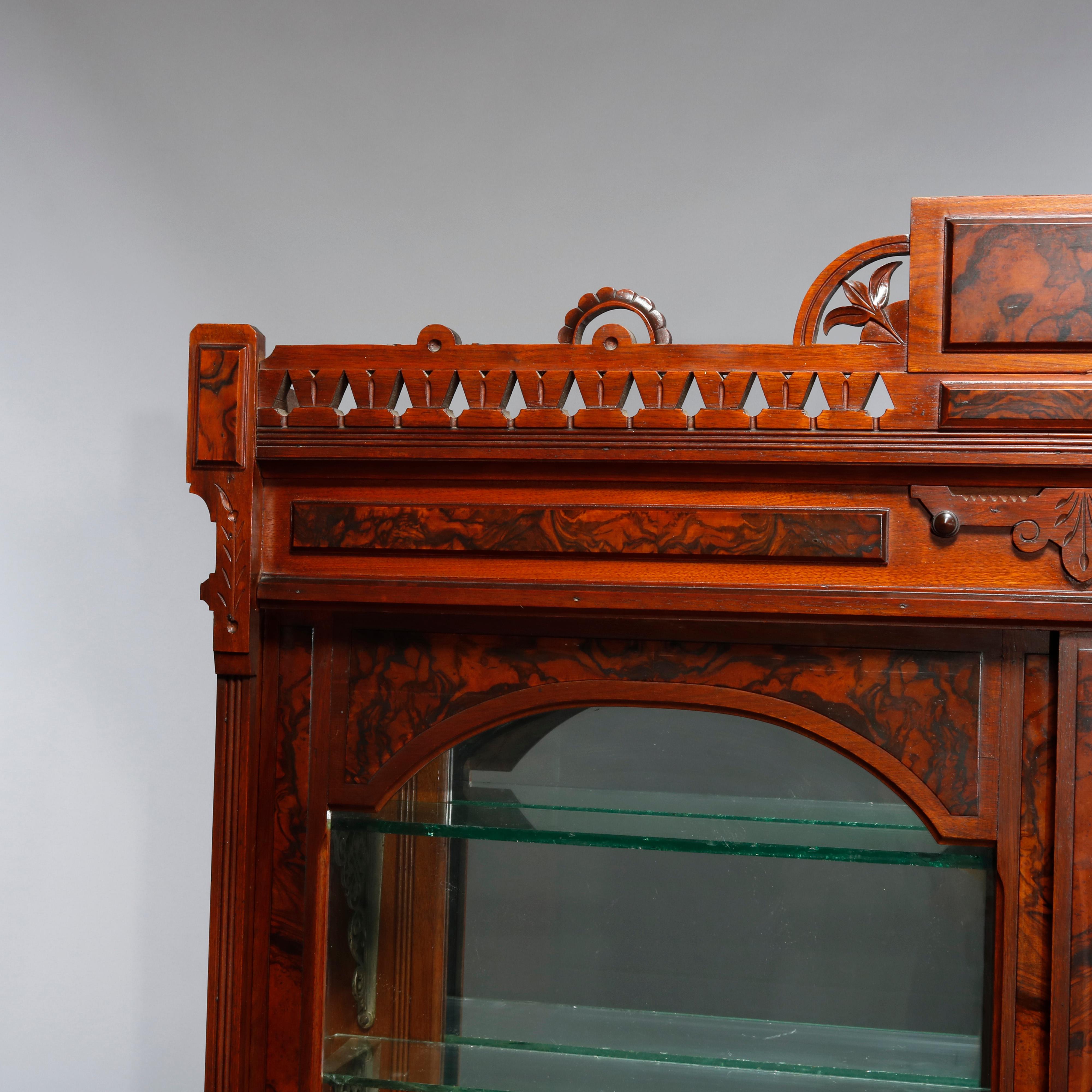 An antique eastlake display case offers walnut construction with pierced rail having central burl crest and flanking carved foliate elements over double doors having arched windows and opening to a shelved and mirrored interior, surmounting lower
