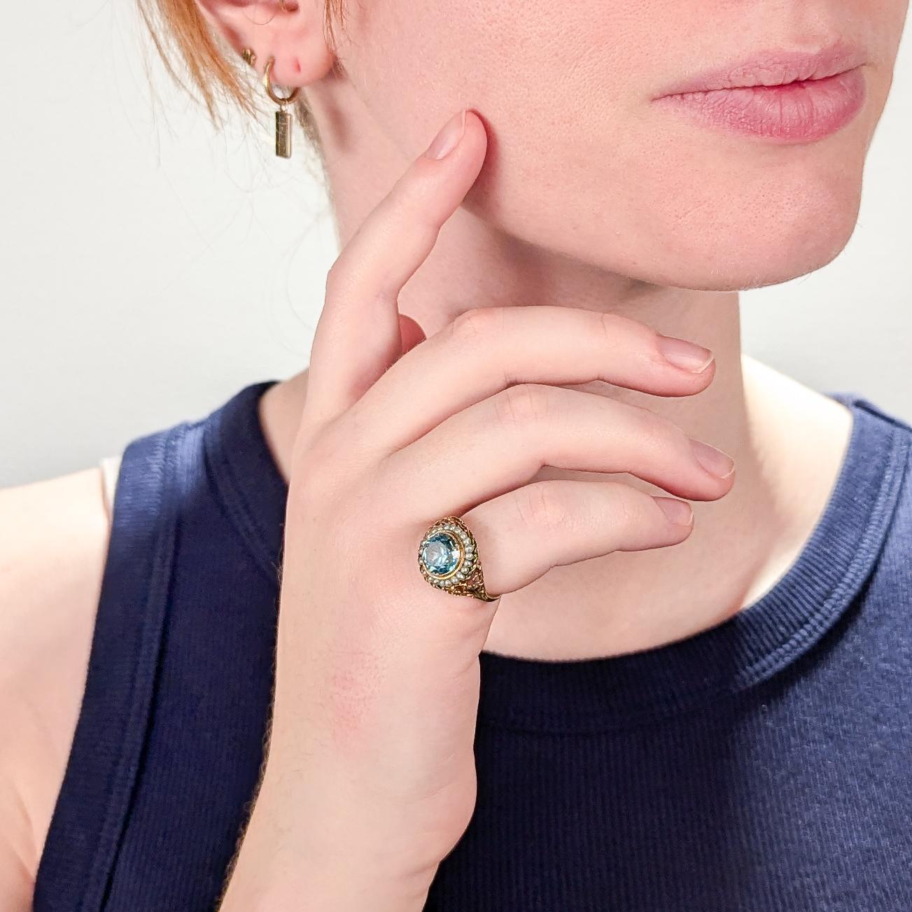 A fine Edwardian signet style ring.

In 14 karat yellow gold.

Bezel-set to the center with a light blue topaz gemstone that is surrounded by a halo of pale blue seed pearls.

Set in an ornate mount with a rope twist edge, with openwork shoulders