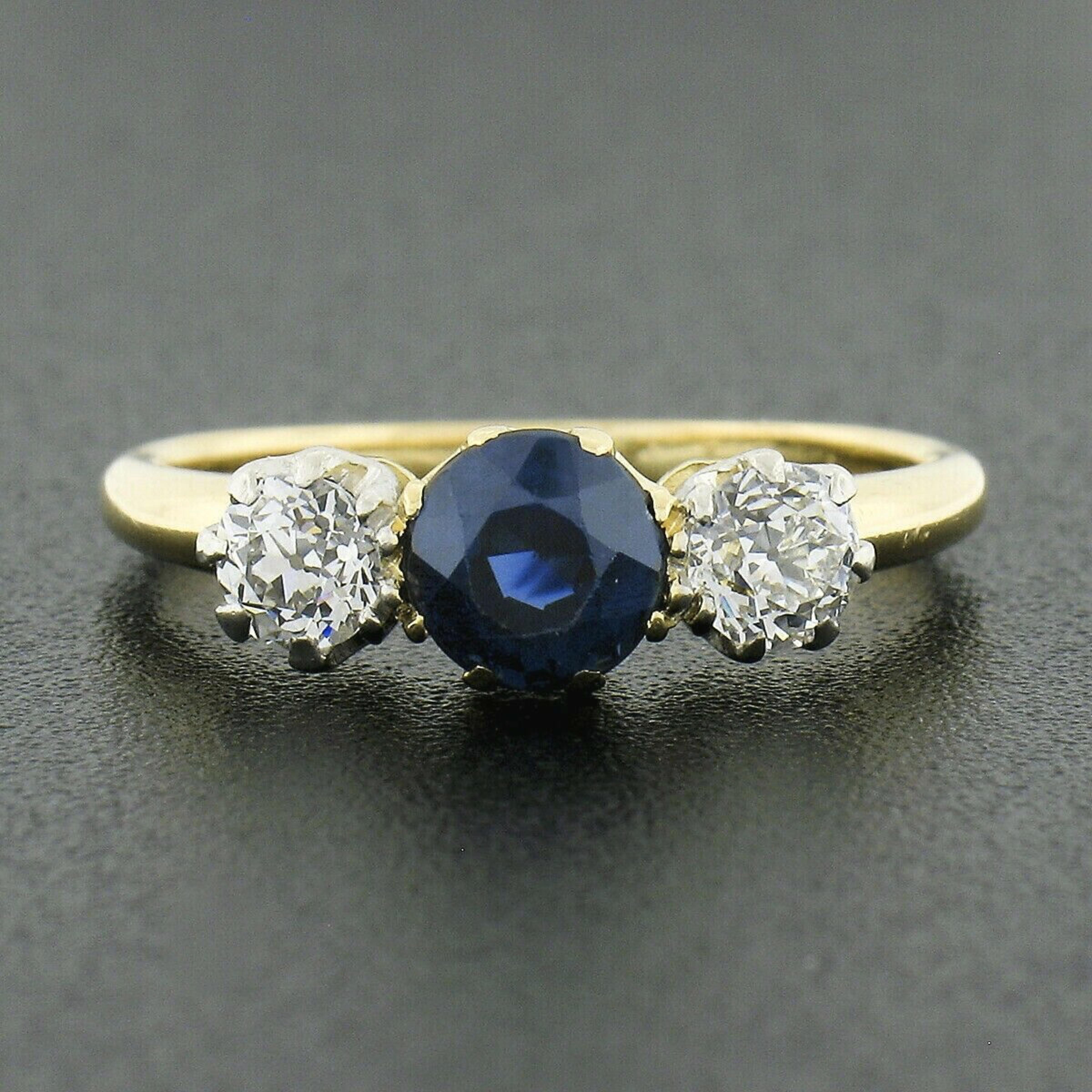 This gorgeous all original antique sapphire and diamond ring was crafted from solid 18k yellow gold during the Edwardian period. It features an elegant three-stone design set at its center with a stunning, older round cut, natural sapphire gemstone