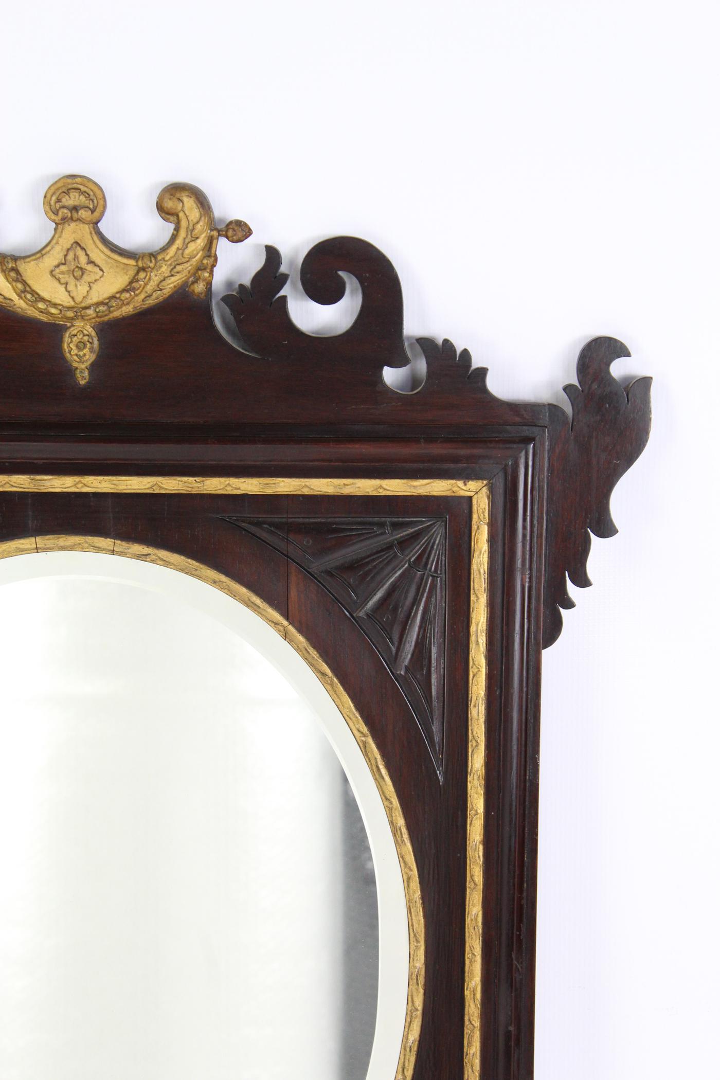 An antique Edwardian mahogany and parcel gilt framed fretwork wall mirror dating from circa 1910. The scroll pediment with a gilt crest and swag, the oval bevelled mirror plate with moulded gilt slip surround and carved spandrels to each corner.