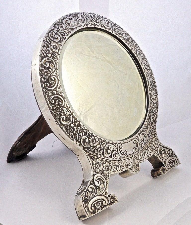 Wonderful antique Edwardian round bevelled edge table mirror, featuring classic sterling silver all over repousse decoration with flowers, leaves and scrolls. There is a plain centre cartouche to the front, and a hinged easel to the back. Mounted on