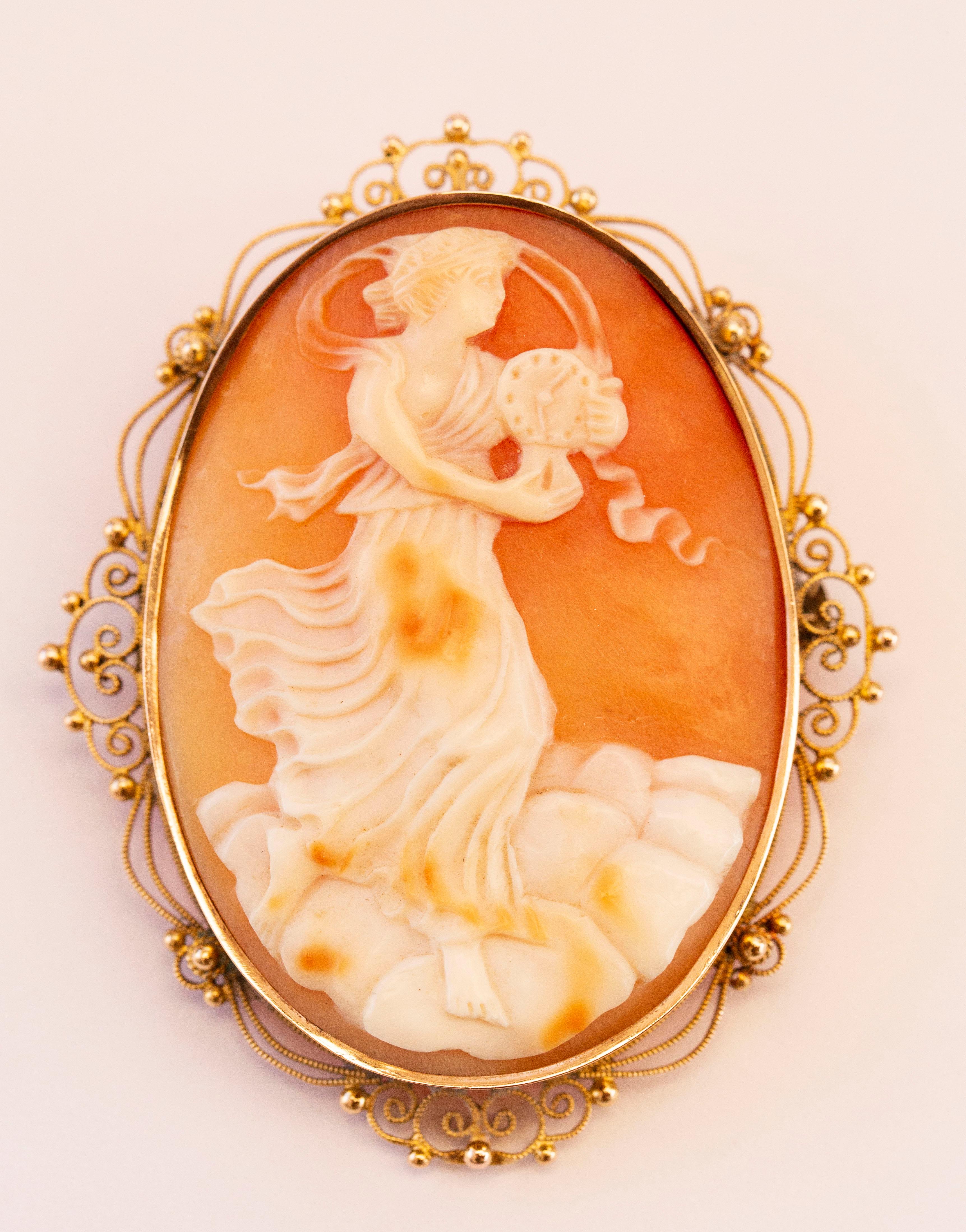 An antique Edwardian (1901-1920) hand carved shell cameo brooch in 14 karat yellow gold filigree frame. The cameo depicts a woman in a long dress with a clock in her hands. The carving is precisely made with refined details and therefore features