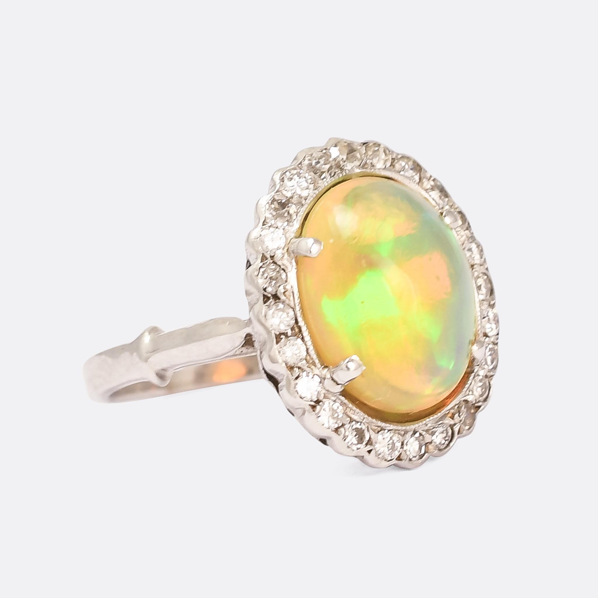 An incredible antique Jelly Opal cluster ring in 18 karat white gold and platinum. The principal stone is a deep cabochon cut, displaying jelly-like splashes of colour - greens, yellows, oranges, and pinks - as it moves through the light. Opals are