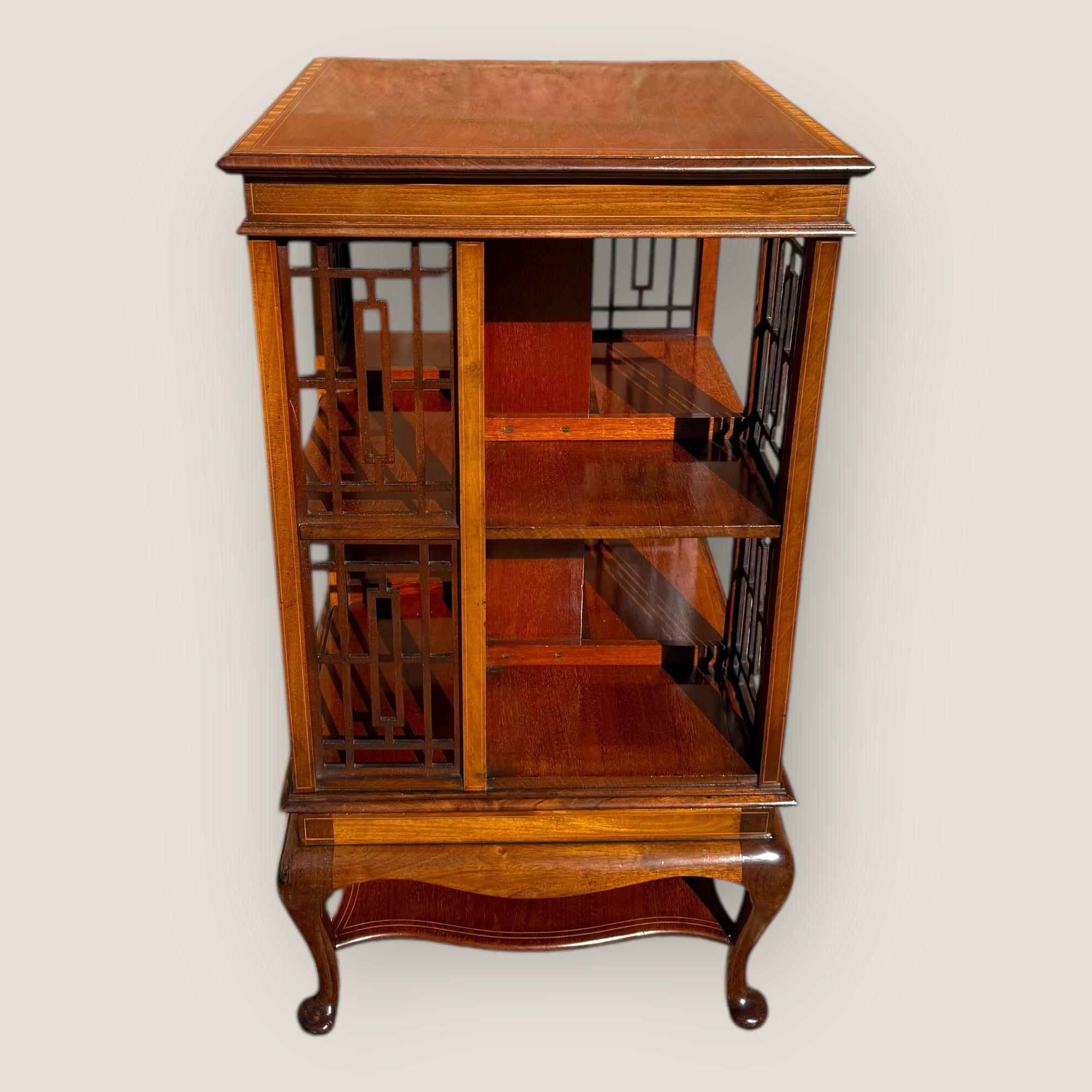 An antique Edwardian mahogany revolving bookcase, circa 1901–1910, inlaid with satinwood and standing on four cabriole legs. The two-tier bookcase is divided into four sections on each level, revolving smoothly to allow convenient access to books