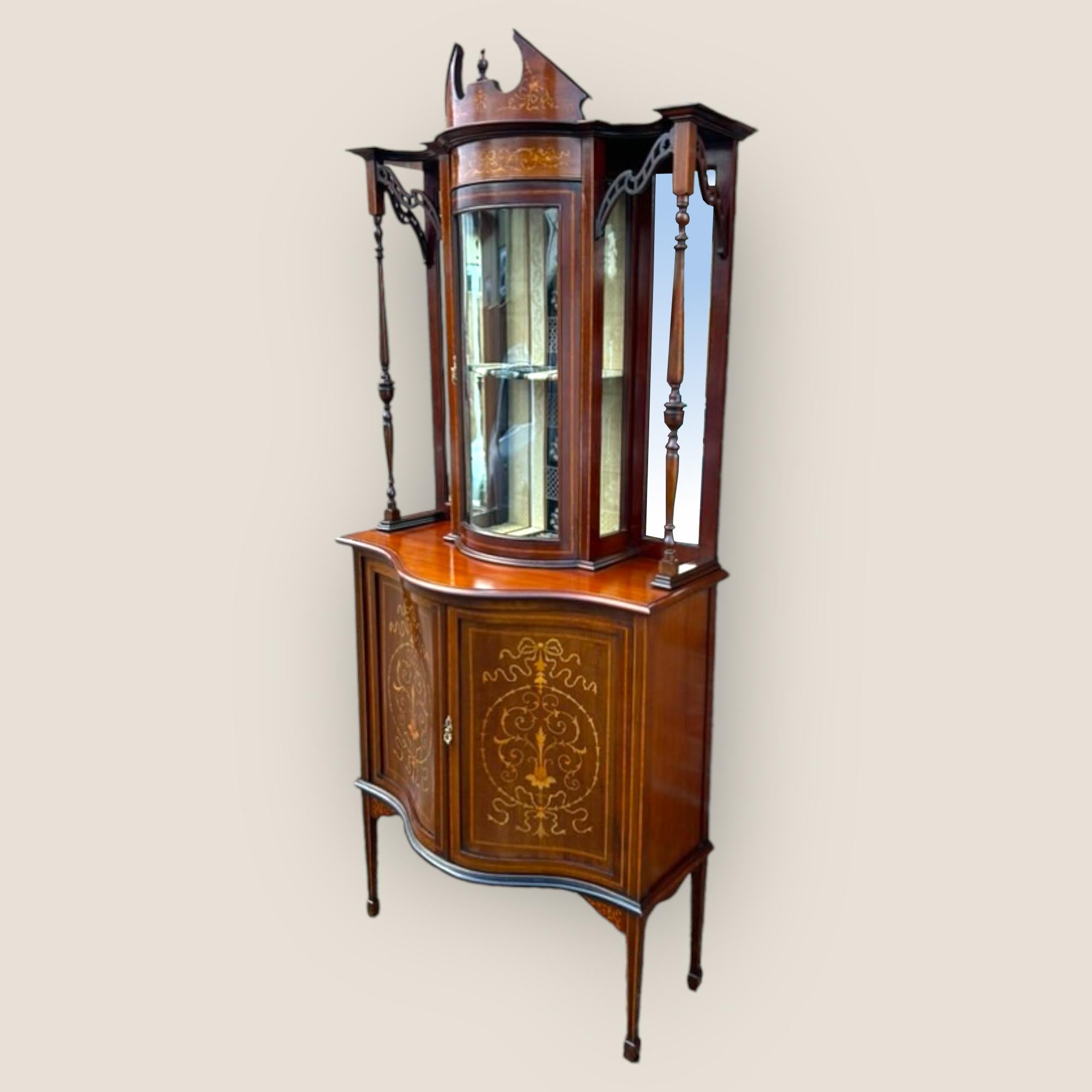 Un mobile da esposizione edoardiano (1901-1910) in mogano e legno satinato intarsiato. Una vetrina centrale in vetro bombato che racchiude due ripiani e un interno a motivi dorati. Ai lati del centro ci sono due specchi smussati e due colonne di