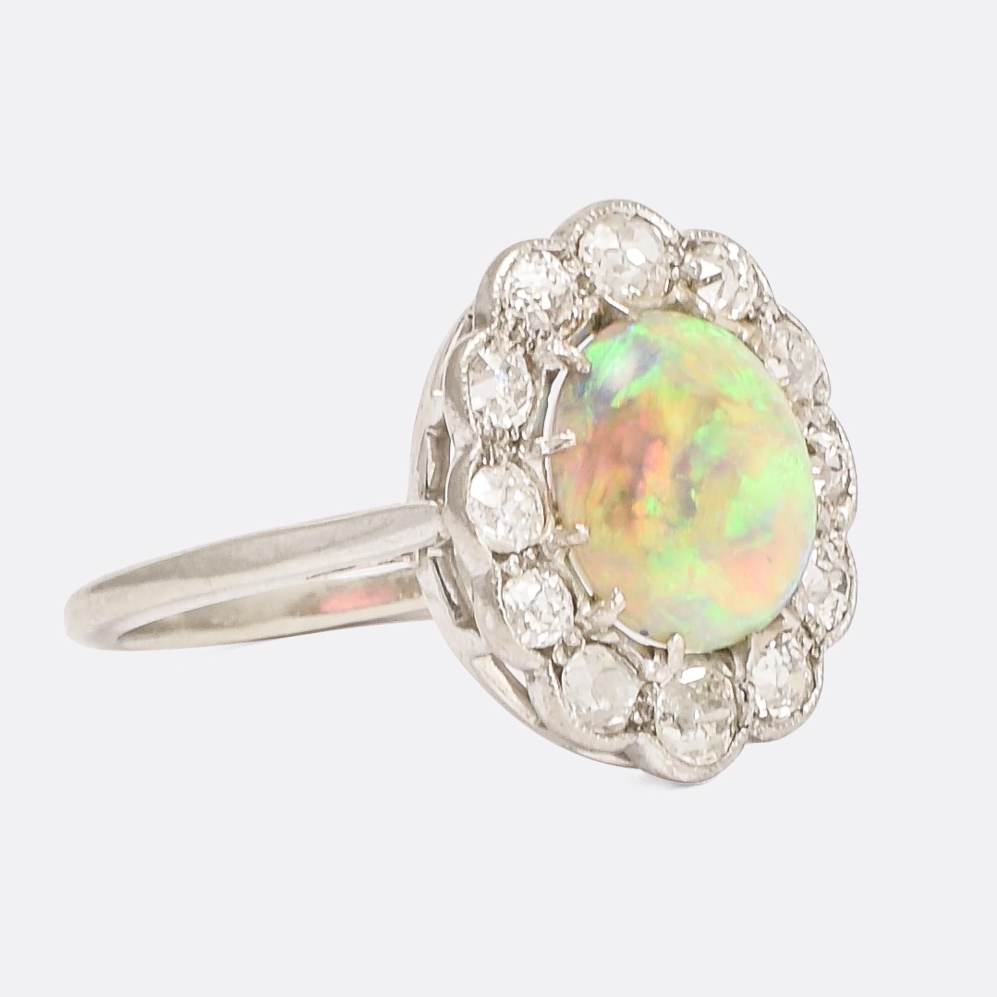 A stunning opal and diamond cluster ring dating from the early 20th Century. The main event is a fiery white opal, displaying vibrant greens, blues and yellows with an orange underglow. It's surrounded by a halo of old mine cut diamonds - totalling