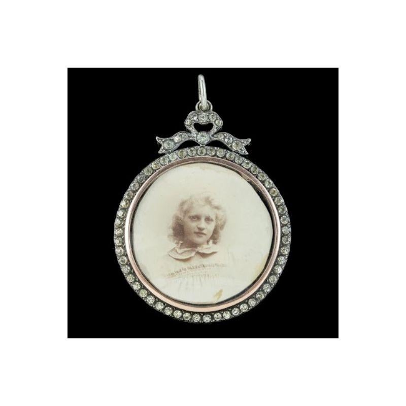 A wonderful Antique Edwardian locket featuring a large glass window at its heart with an original photograph of a young lady stored safely behind. This is haloed by an array of glistening white Paste Stones and topped with a bow. 

It’s fashioned in