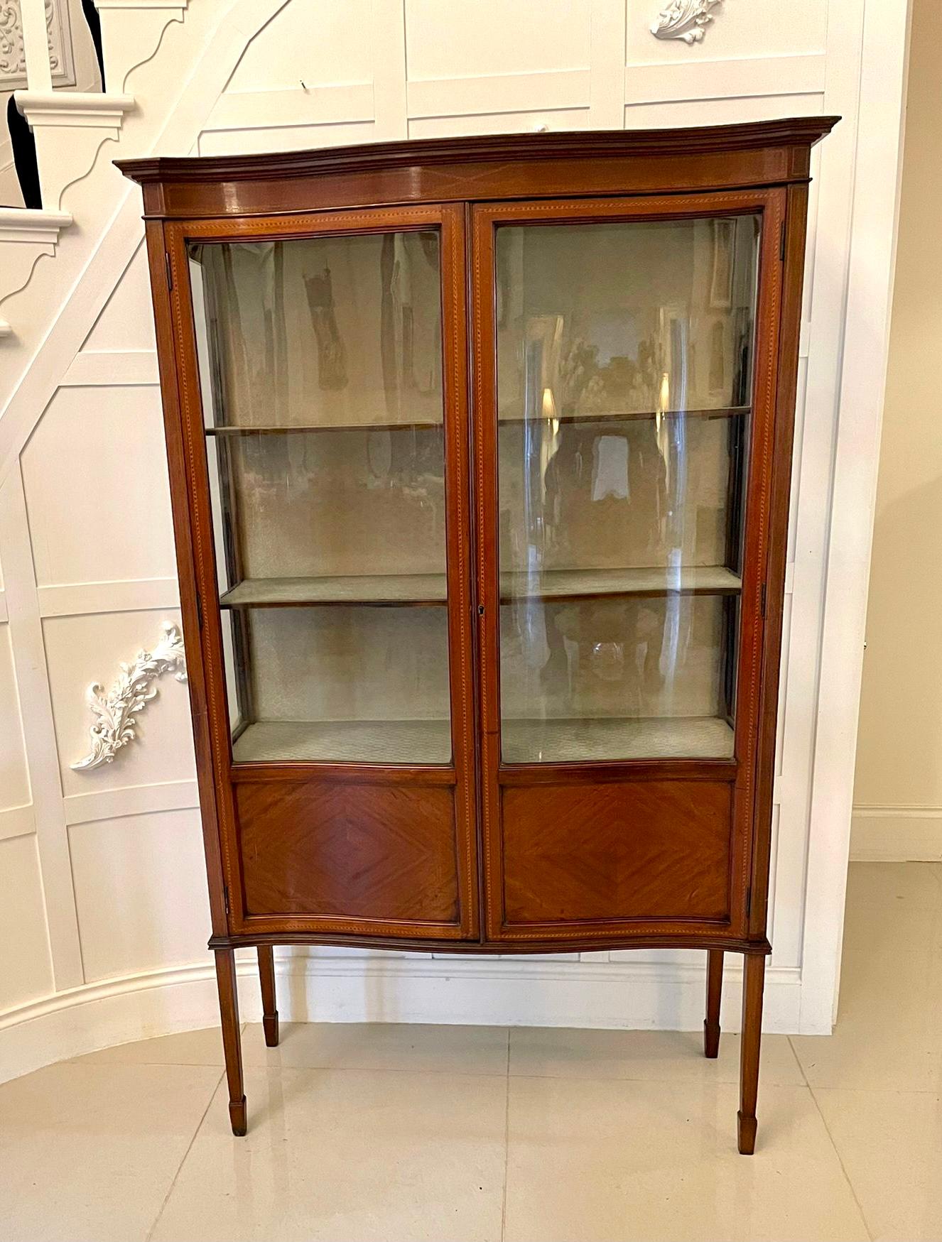 Antique Edwardian quality mahogany inlaid serpentine front display cabinet having a quality mahogany shaped cornice, mahogany inlaid frieze above a pair of pretty serpentine shaped glazed and mahogany inlaid doors opening to reveal three fabric