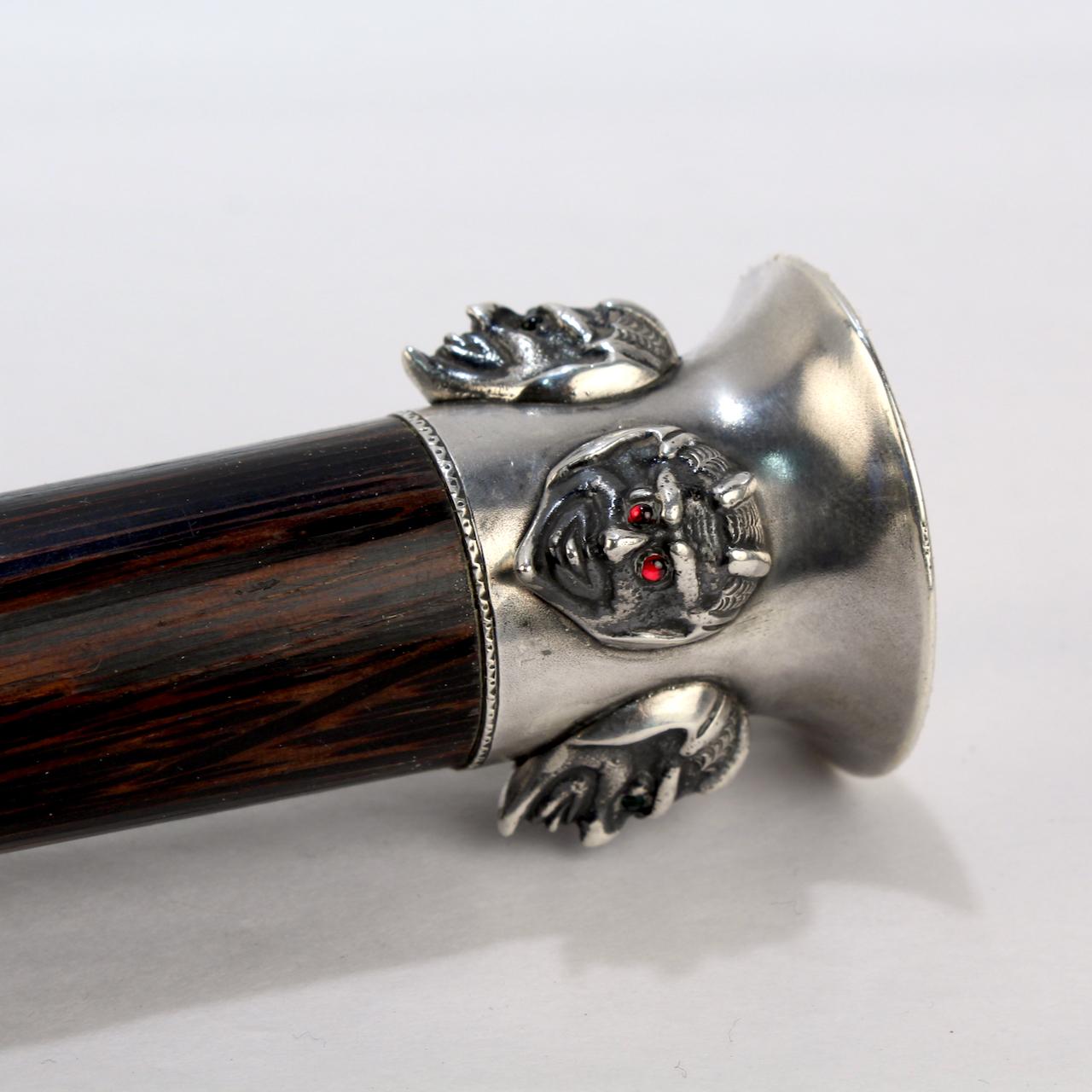 A fine Edwardian cane or walking stick.

With a silver cane handle that has 4 devil's heads or masks around its circumference. The faces have alternately tiny ruby red and emerald green glass cabochon eyes. 

The top of the handle is engraved