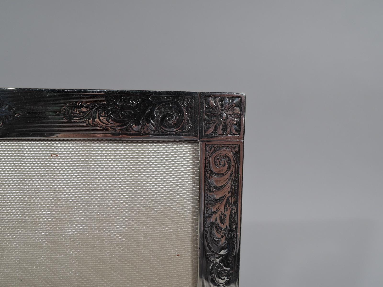 Edwardian sterling silver picture frame. Made by Birks in Canada, circa 1910. Rectangular window and flat surround with wraparound engine-turned lines and chased leafy scrolls and corner paterae. At top shaped rectilinear cartouche (vacant). With