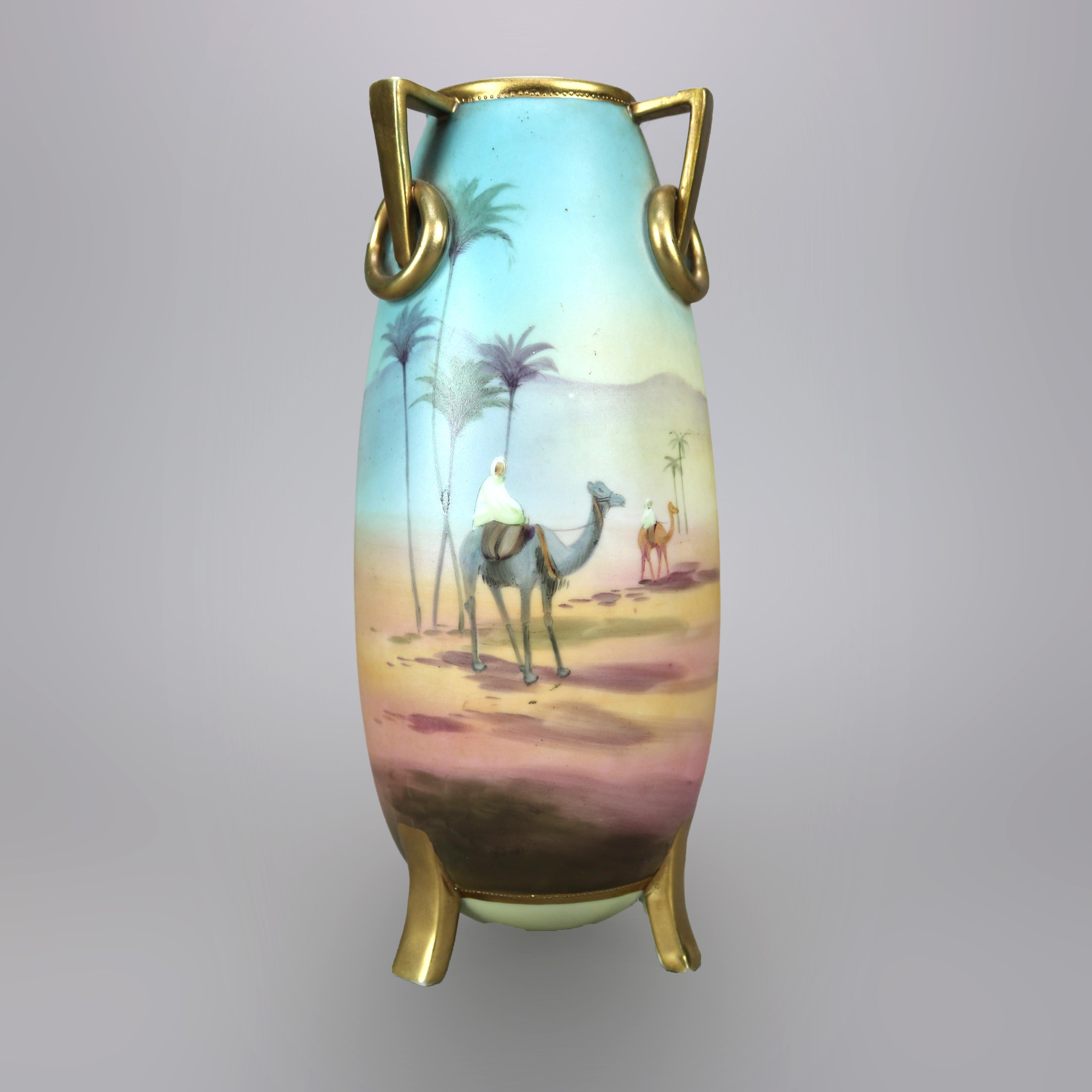 An antique Egyptian Revival Nippon vase offers porcelain construction with all-around hand painted Persian desert scene having figures and camels, gilt buttress handles having rings and raised on gilt tripod and flared legs, maker stamp on base as