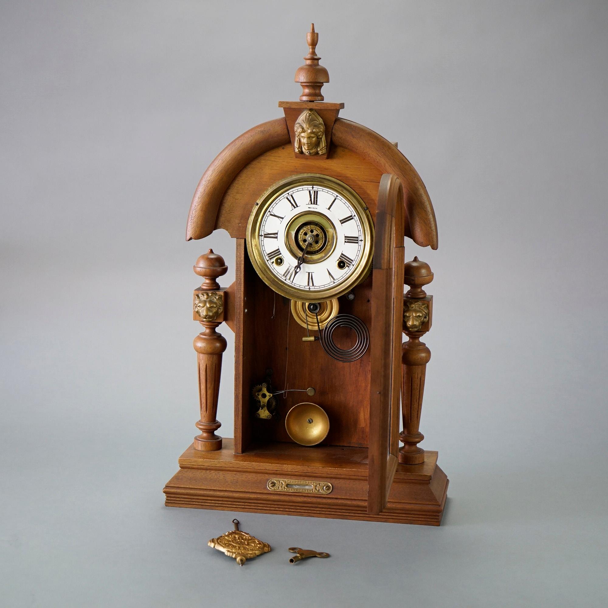 An antique Victorian Egyptian Revival style mantel clock offers carved walnut frame in arched form with urn form finial over cast mount in the form of a female mask, door with reverse painted decoration and flanking turned columns,