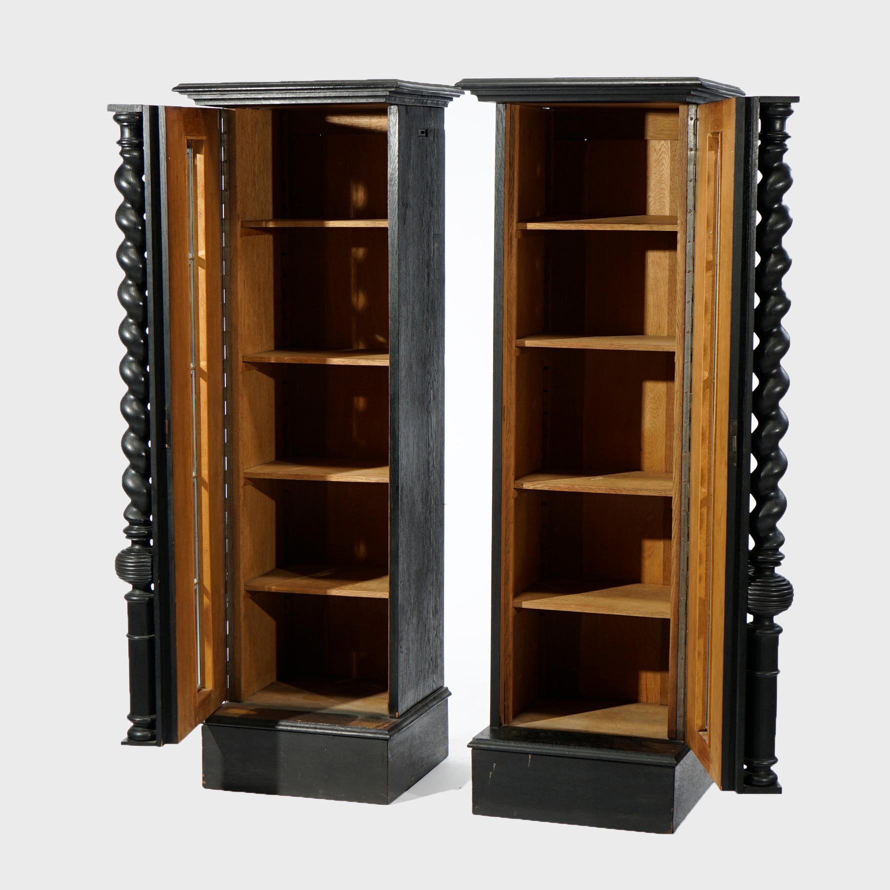 An antique pair of Elizabethan style bookcase cabinets offer ebonized English oak construction in tall narrow form, each having a single door with leaded beveled glass opening to shelved interiors and with flanking barley twist columns,