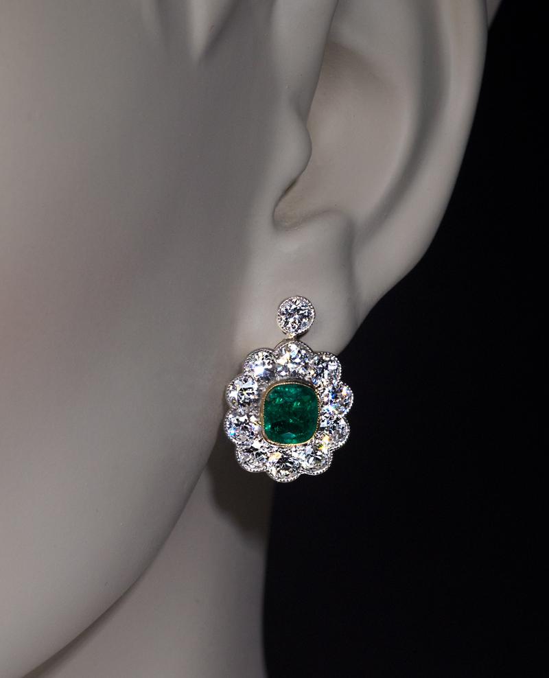 Circa 1910

A pair of antique Edwardian era cluster earrings is crafted in platinum and 14K gold. The earrings are centered with cushion cut emeralds of an excellent deep bluish green color framed by bright white and clean (E-F color, VS1-SI1