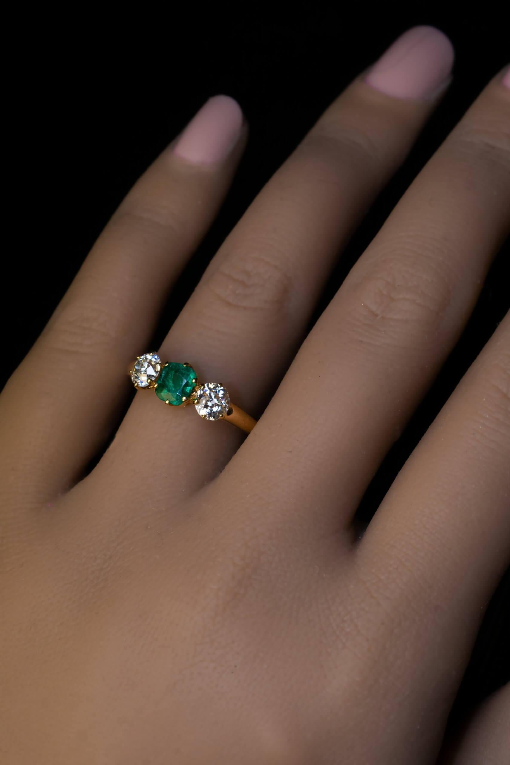 Circa 1890  This classic Victorian era three stone ring is crafted in 14K gold. The ring is centered with a cushion cut emerald of a deep bluish green color (likely of Colombian origin).  The emerald is flanked by two bright white and clean old mine