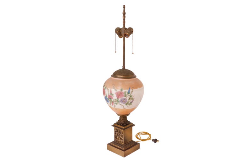 An antique Empire style brass lamp with a hand painted glass globe. A slender central column topped with an urn finial has two sockets that light with brass chain pulls. A blush and white glass globe secured with an acanthus pressed brass cap is