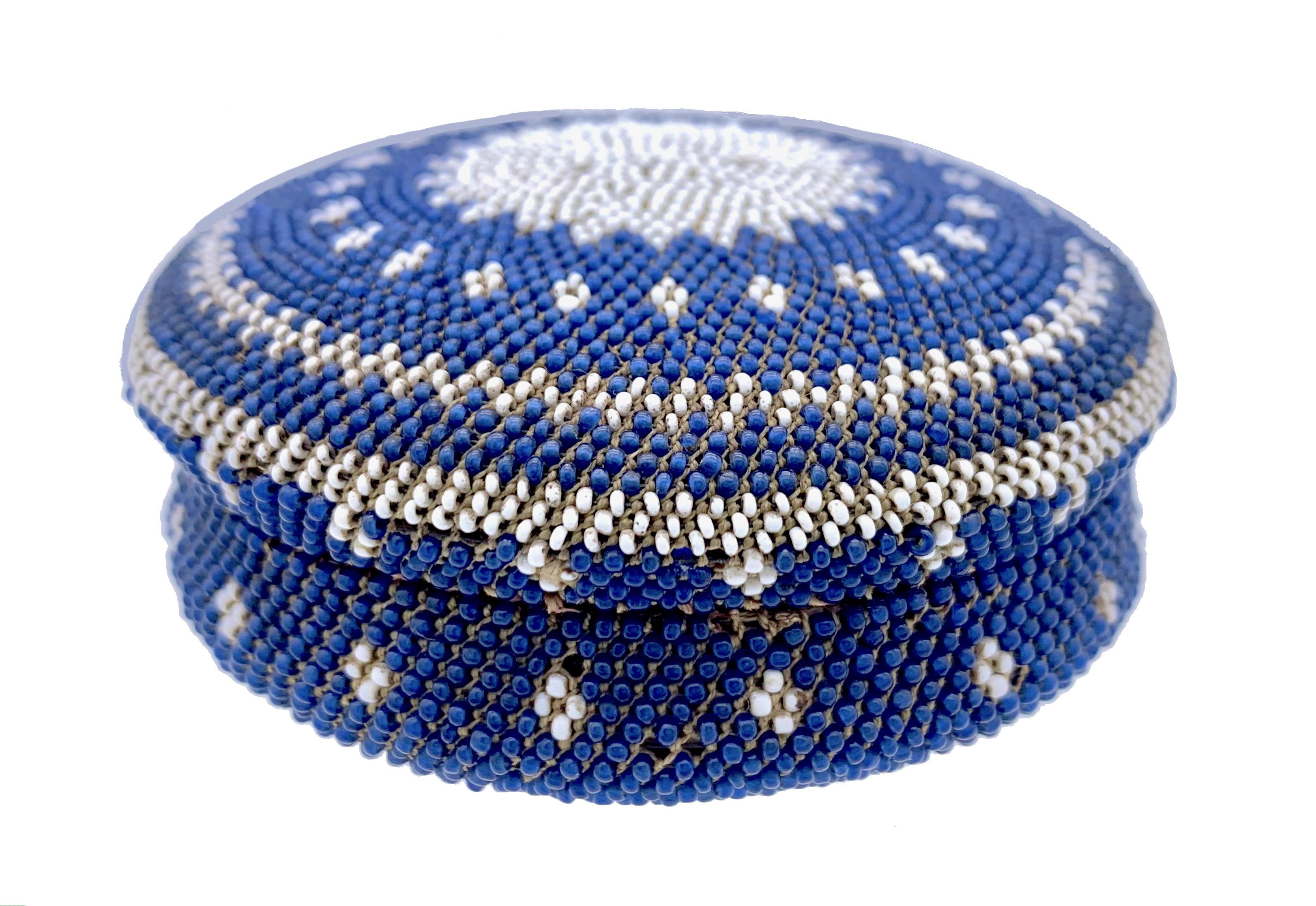 Esta delicada tabaquera se ha fabricado con cuerno prensado y está recubierta por completo de cuentas de vidrio azules y blancas ensartadas en hilo de algodón. La tapa está decorada con un emblema solar, un rombo y un dibujo en zigzag y las palabras