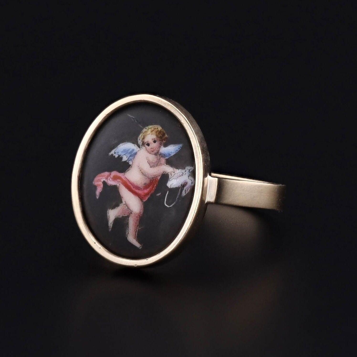 Cette bague en or 14k est ornée d'un chérubin émaillé tenant une minuscule colombe. La face de la bague était à l'origine une épingle ancienne (vers 1890) que nous avons transformée en retirant l'épingle de son support d'origine et en ajoutant un