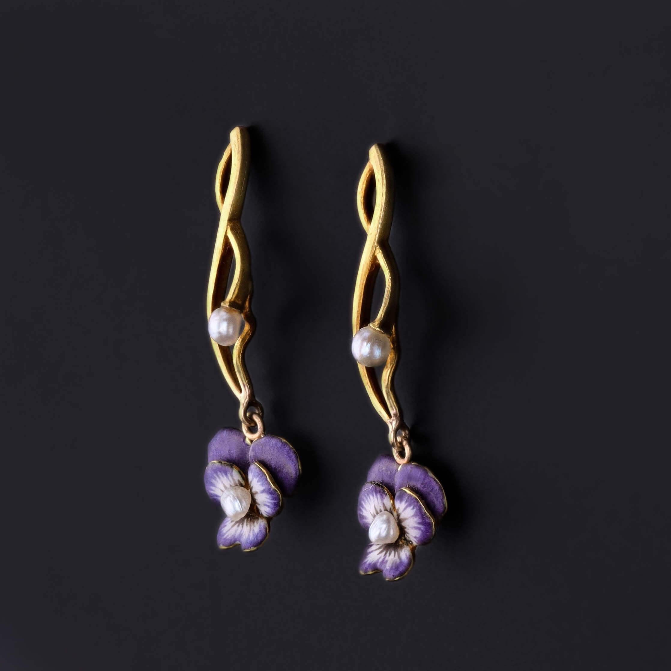 Ces charmantes boucles d'oreilles anciennes présentent des pensées en émail violet et en perles suspendues à des pendants en or 14 carats, mêlant l'art floral délicat à un mouvement gracieux. Conçus à l'origine comme d'anciennes épingles à dentelle