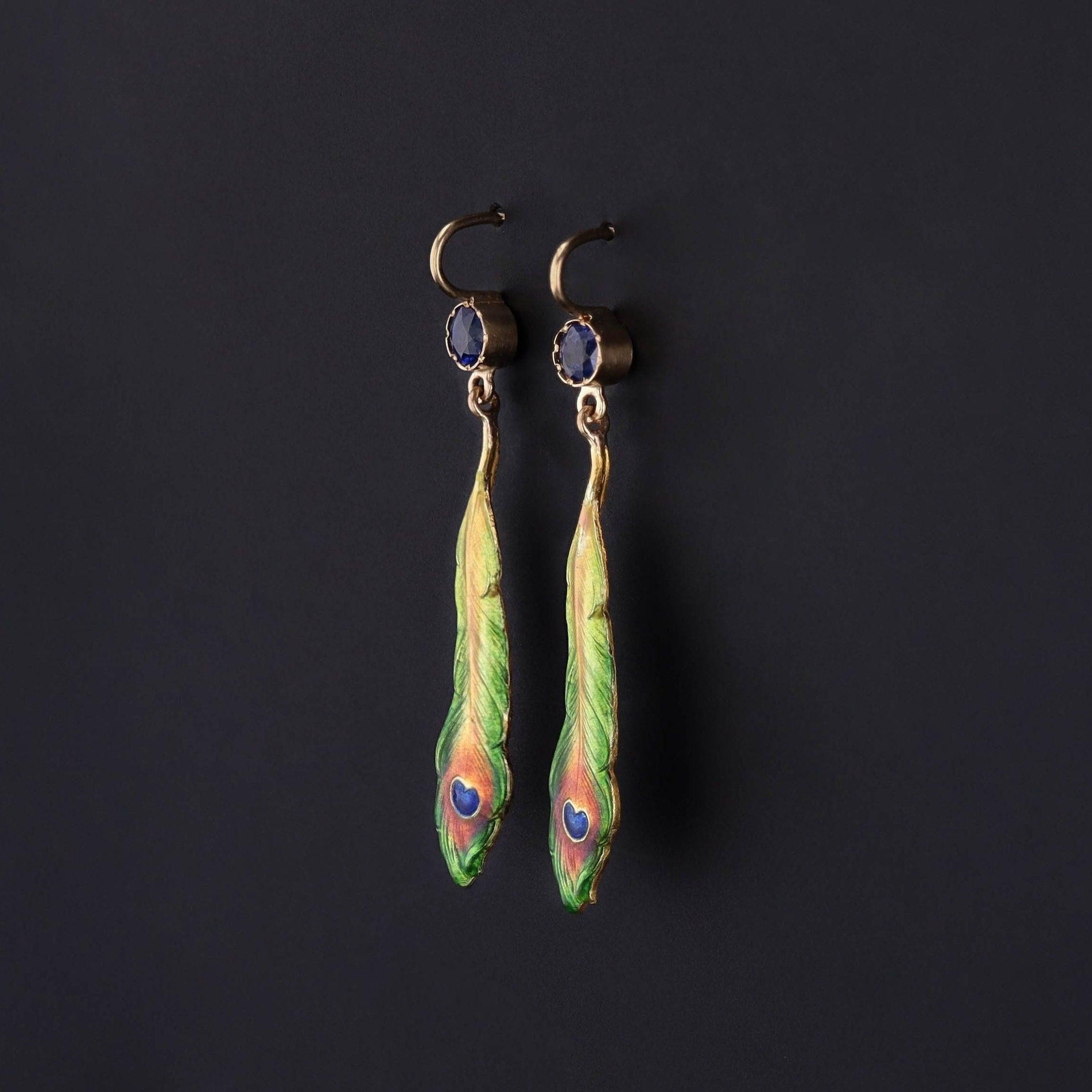 These earrings were created from antique pins (circa 1910) and feature peacock feathers of 14k gold, adorned in enamel and dangling beneath sapphire surmounts.

Each earring measures 1.6 inches from the top of the ear wire to bottom by 0.3 inches