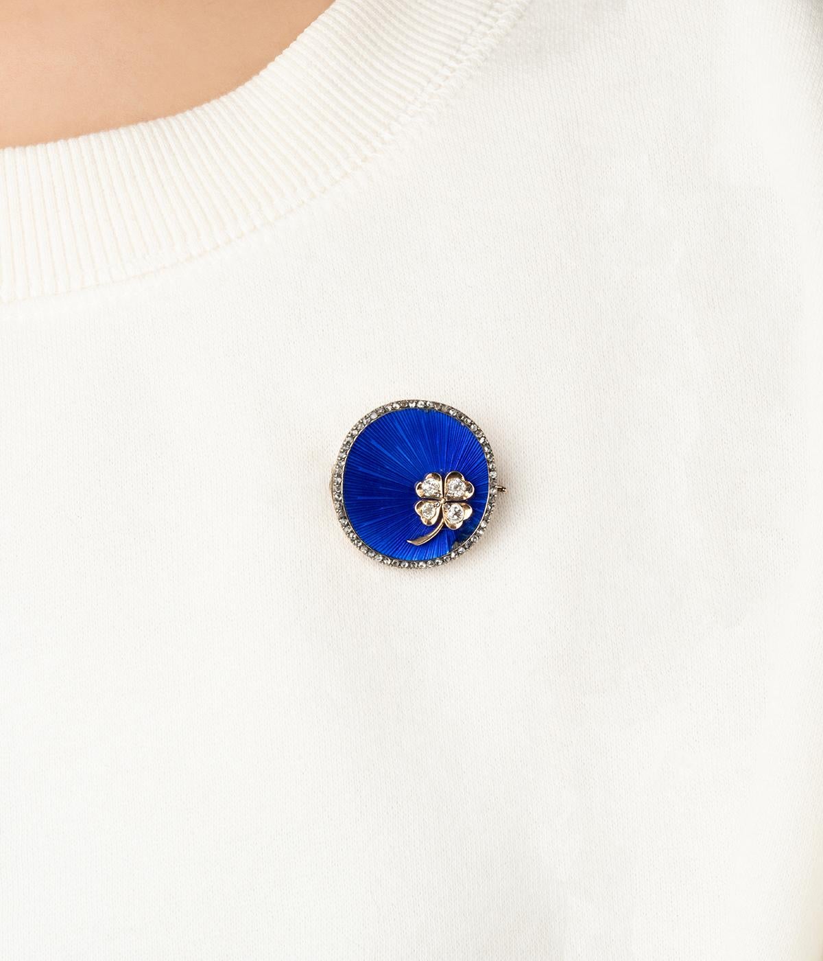 Broche ancienne en or 14 carats (585 millièmes) et argent du début du 20ème siècle. De forme ronde, elle est entourée d'une ligne de diamants taille rose. Son fond est rehaussé d'émail bleu nuit rayonnant et d’un trèfle serti de 4 diamants taille