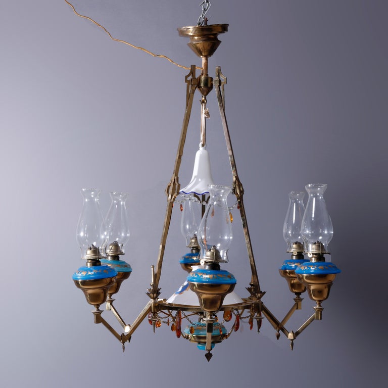 An antique English Aesthetic partially electrified oil lamp chandelier offers cast brass angular frame with central oil lamp electrified and having hand painted cameo shade, six arms extend and terminate in oil lamps having gilt decorated blue