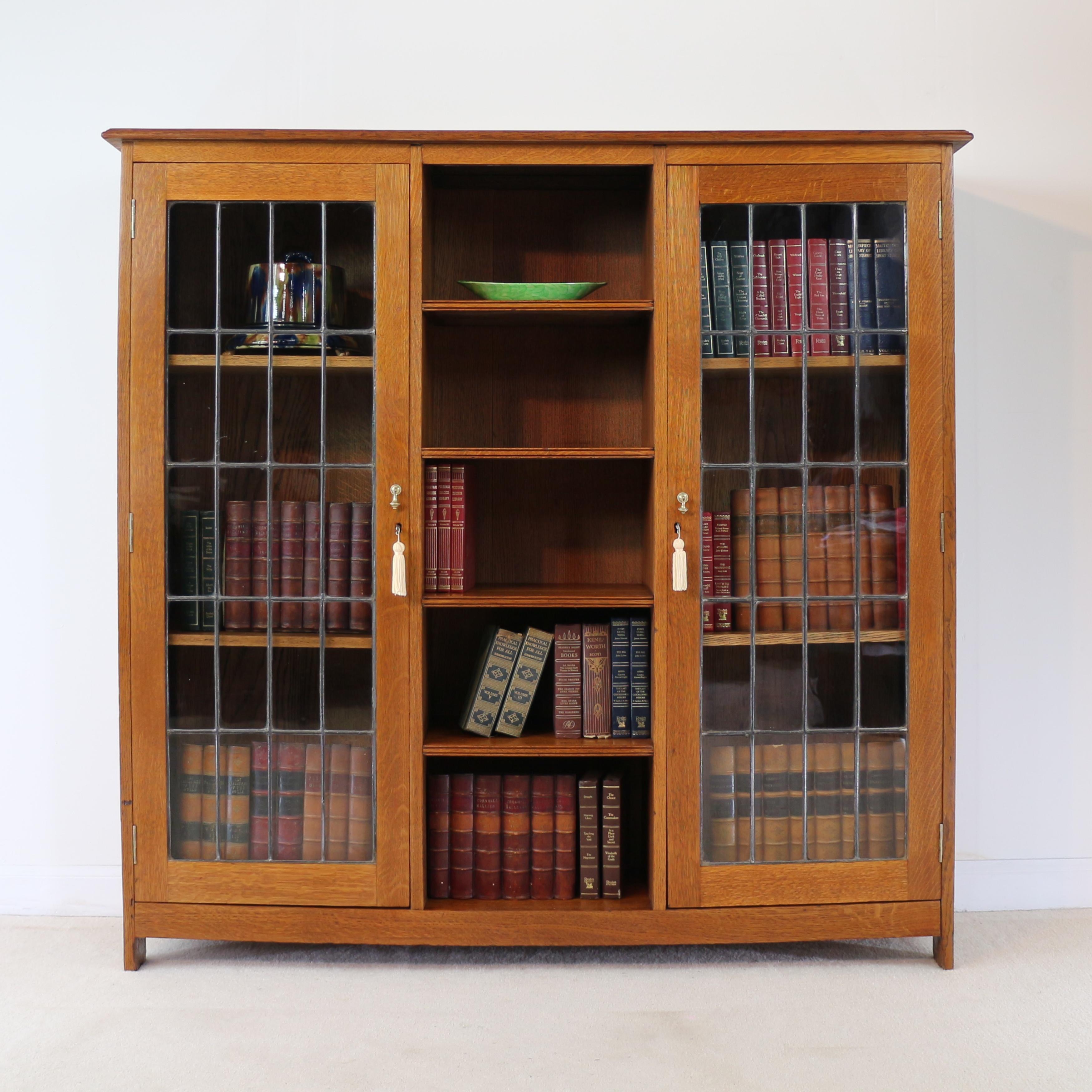An attractive Arts & Crafts oak bookcase in golden quarter-sawn oak, attributed to Liberty’s of London and dating to circa 1900. Featuring two leaded glass doors each enclosing two adjustable shelves, and open shelves to the centre, with brass drop