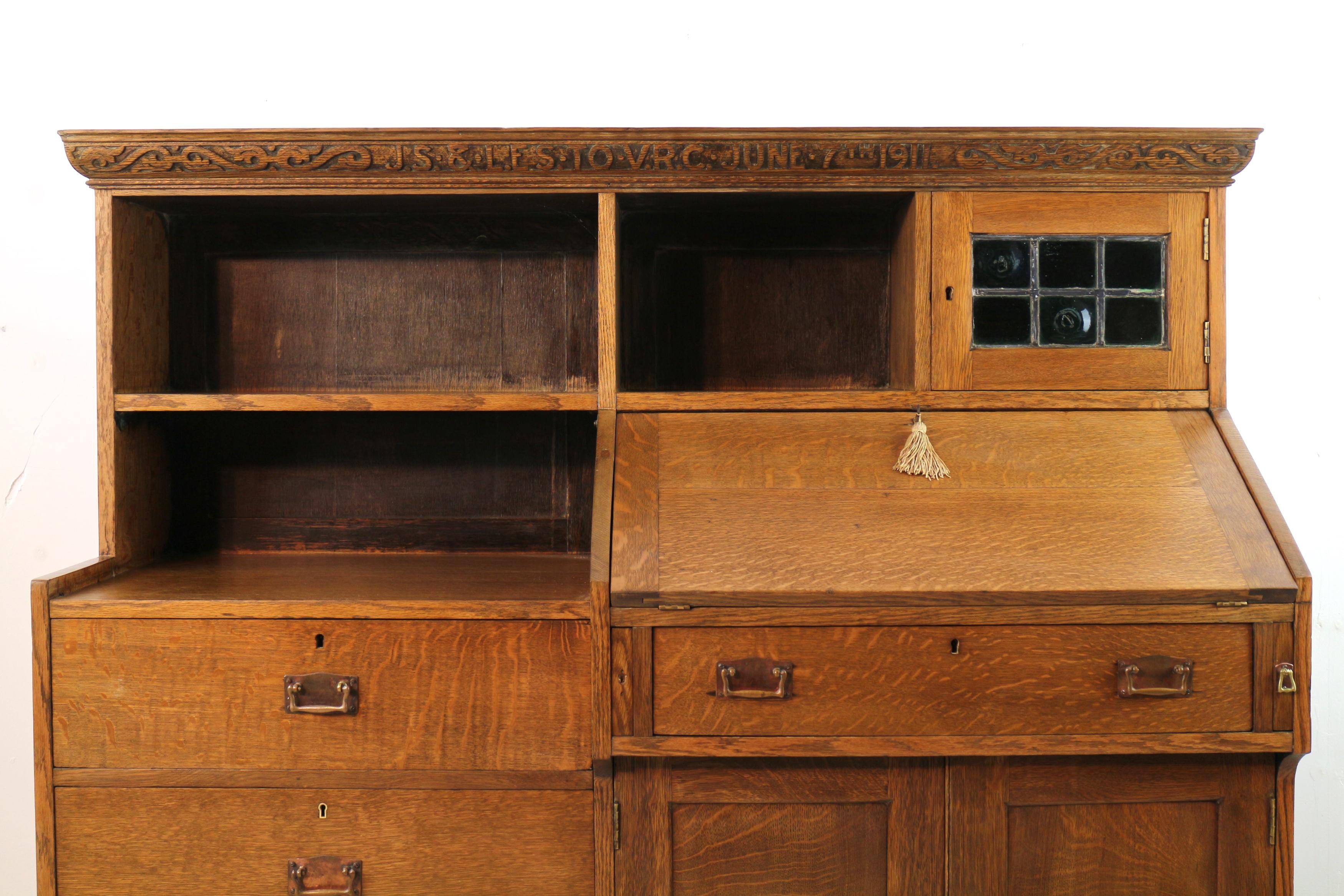 An attractive Arts & Crafts oak motto style bureau bookcase in golden quarter-sawn oak firmly attributed to Liberty & Co. of London. Featuring a moulded carved cornice with inscription ‘J.S. & L.E.S. TO V.R.C. JUNE 7TH 1911’ above a leaded green