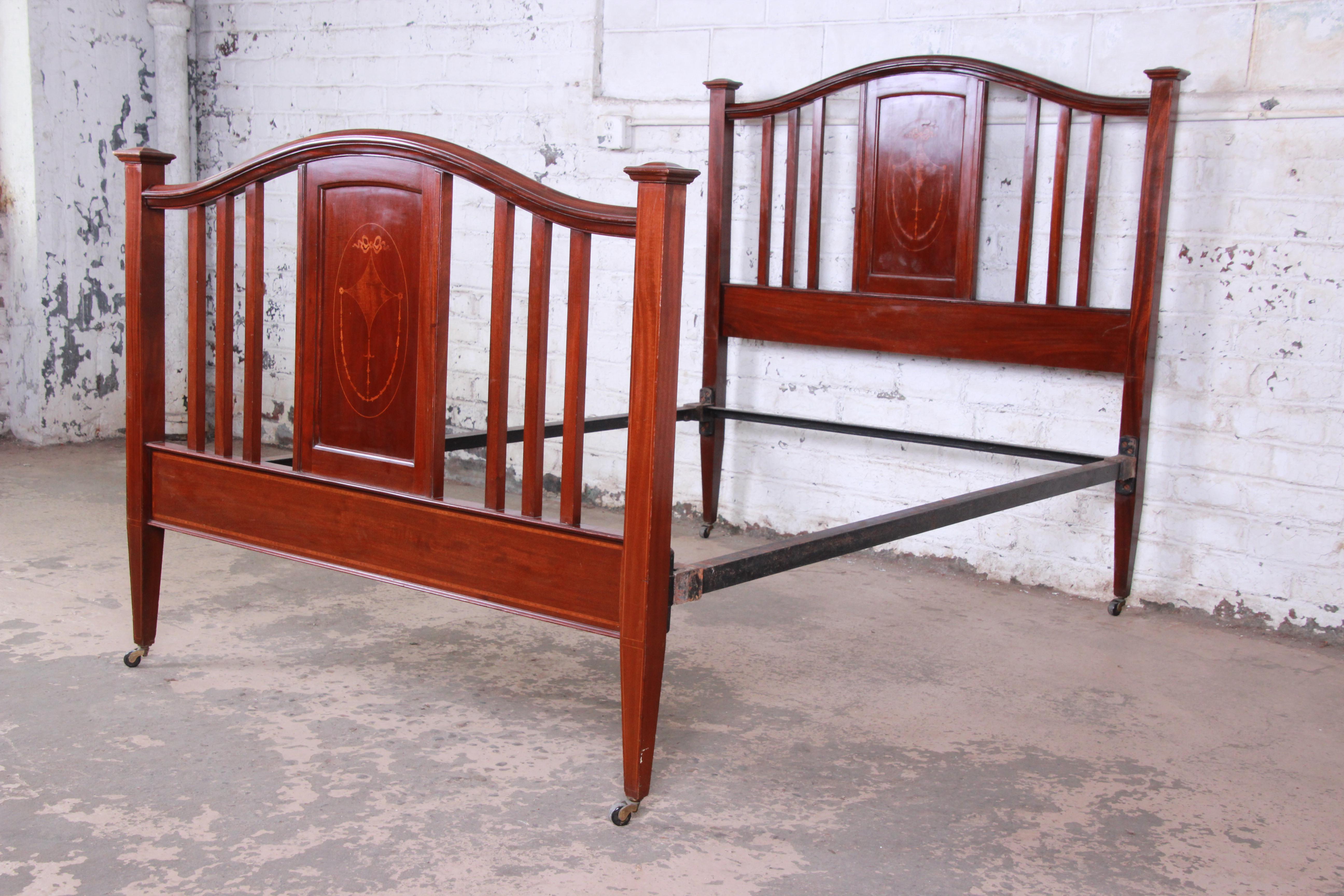 A gorgeous antique English Arts & Crafts inlaid mahogany bed frame. The bed features solid mahogany construction with beautiful inlaid details on both the headboard and footboard. The heavy iron side rails can support either a full or queen size