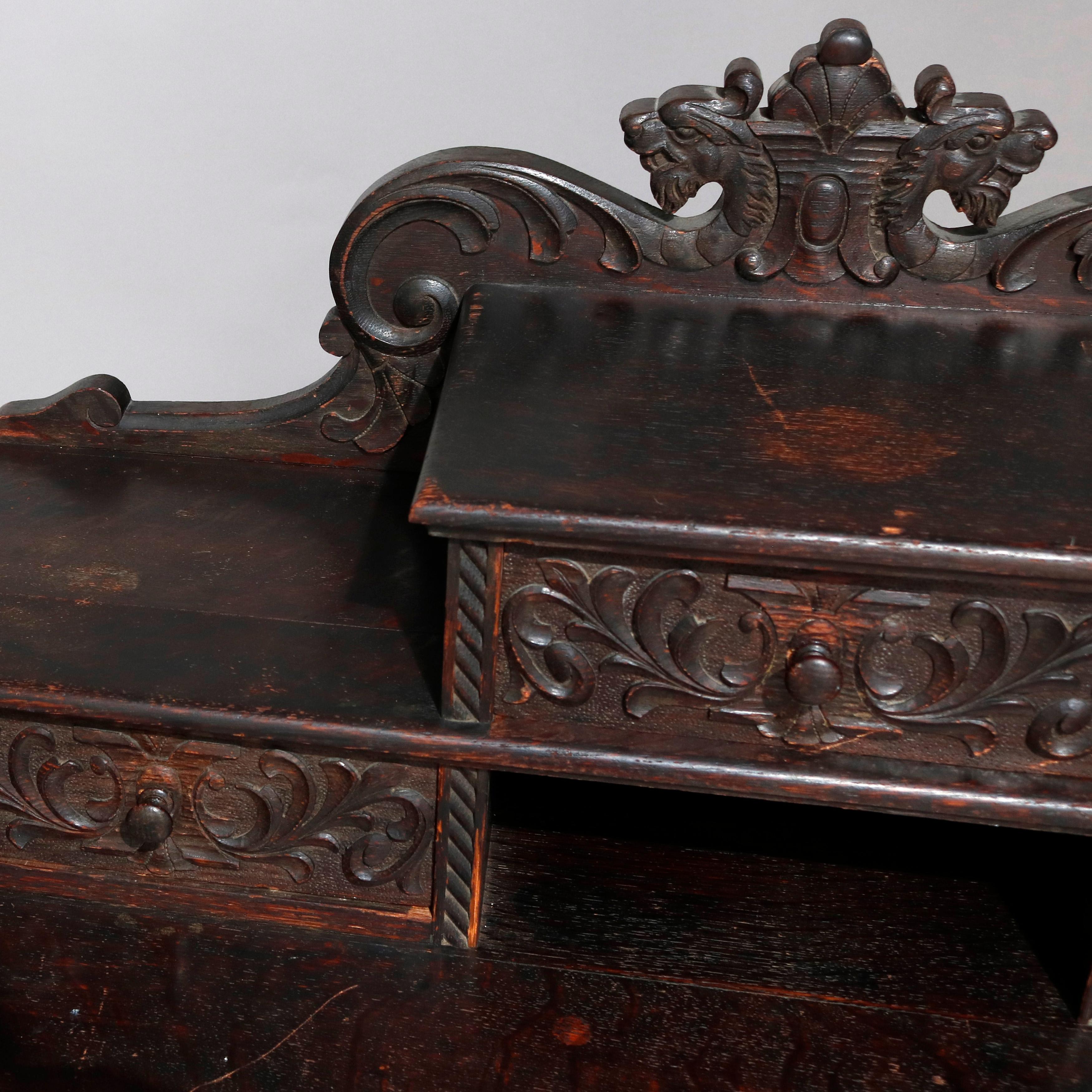 An antique English Baroque plantation desk offers heavily carved oak construction with backsplash having shield form crest and flanking foliate scrolls surmounting stepped tower having three foliate carved drawers surrounding central pigeon hole and