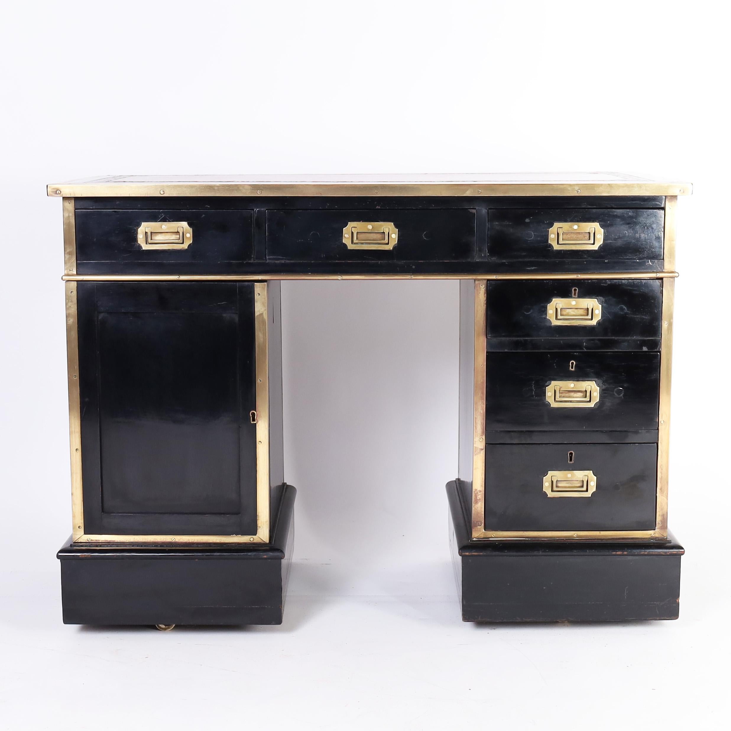 Chic antique English campaign desk crafted in hardwoods having a tooled red leather top over an elegant black lacquer two pedestal form with six drawers and a cabinet bound with campaign brass and set on block feet raised on casters.