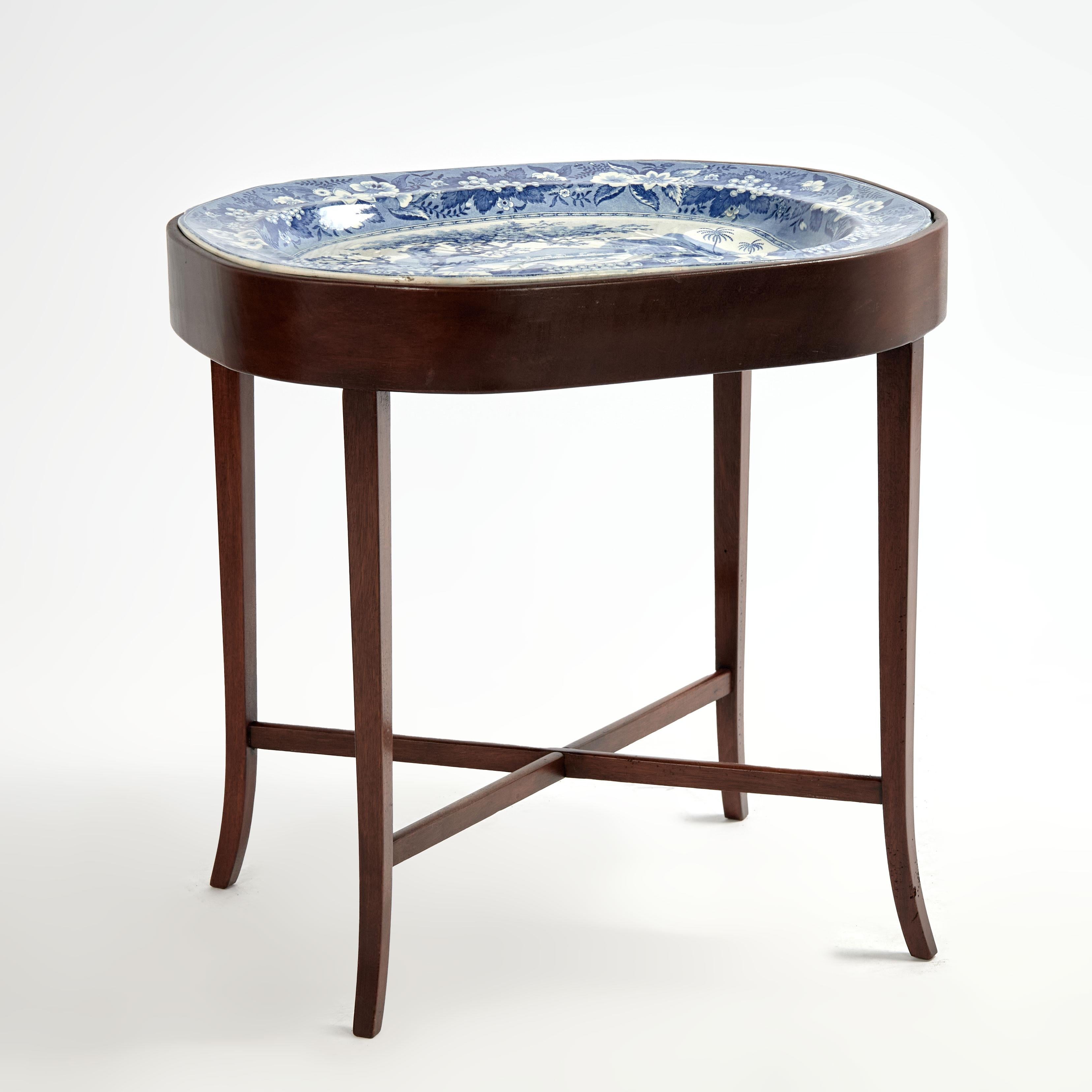 Vassoio inglese del XIX secolo in bianco e blu, decorato con alberi, barche e figure all'interno di un bordo floreale, presentato in un supporto in mogano personalizzato più recente con le classiche gambe strombate e le barelle incrociate. 